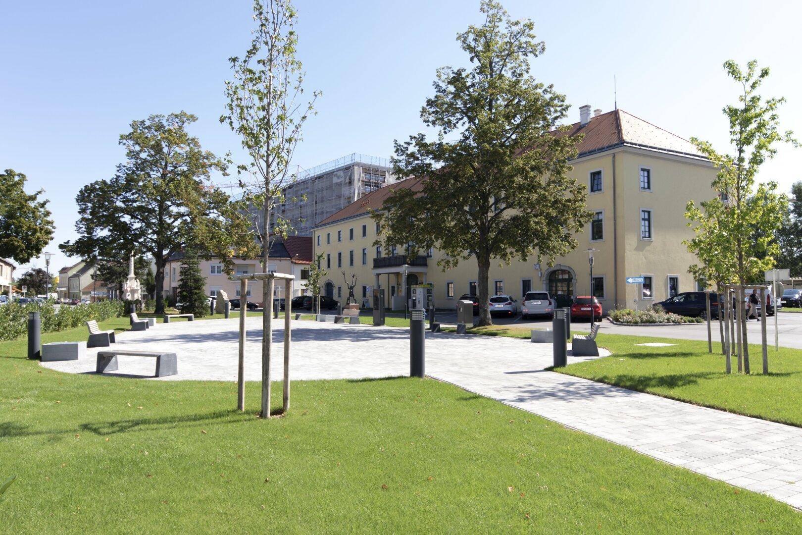 Öffentlicher Platz mit Grünflächen, Bänken und einem historischen Gebäude im Hintergrund.