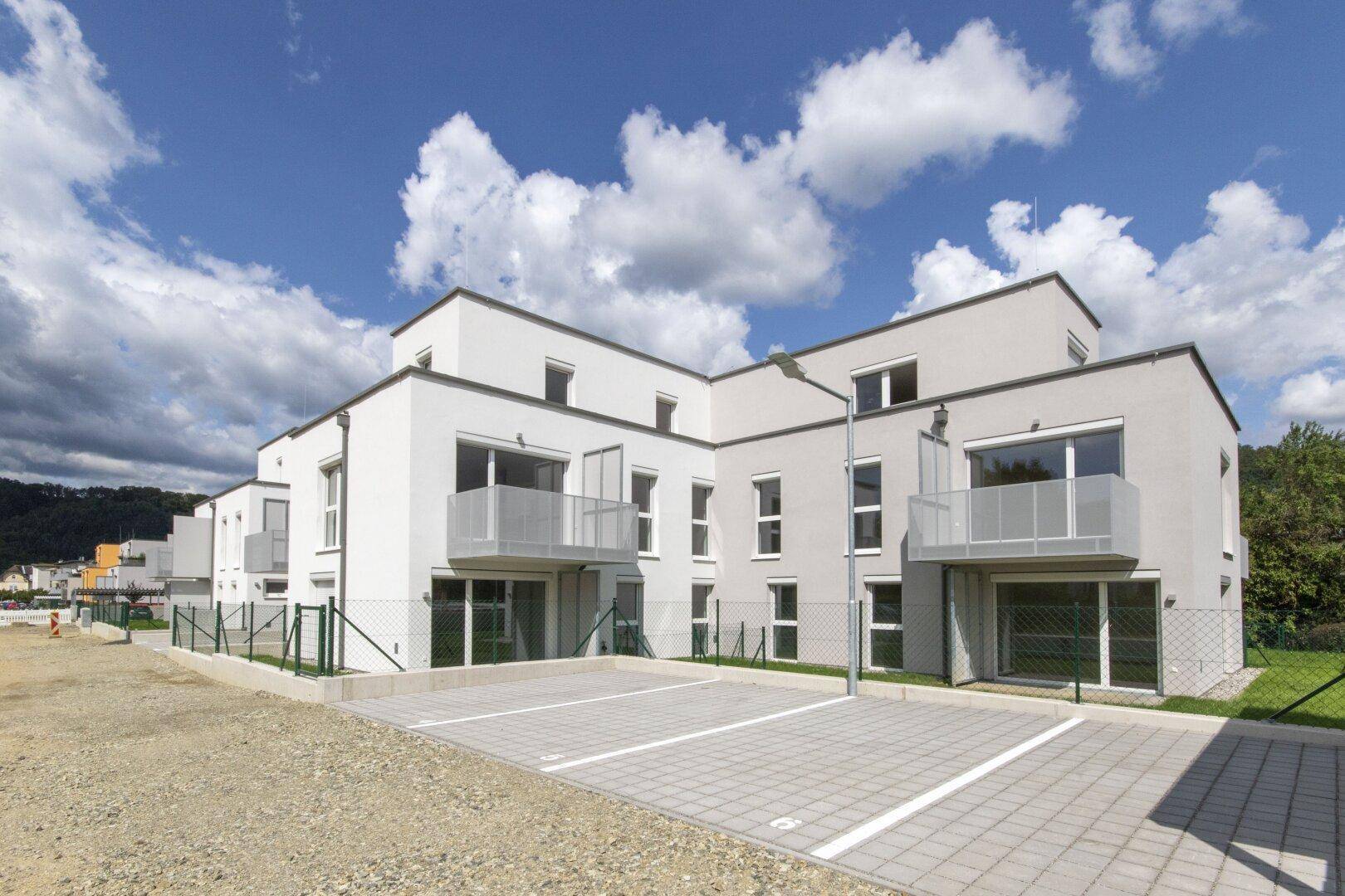 Modernes Mehrfamilienhaus mit weißer Fassade, Balkonen und Außenparkplätzen unter blauem Himmel.