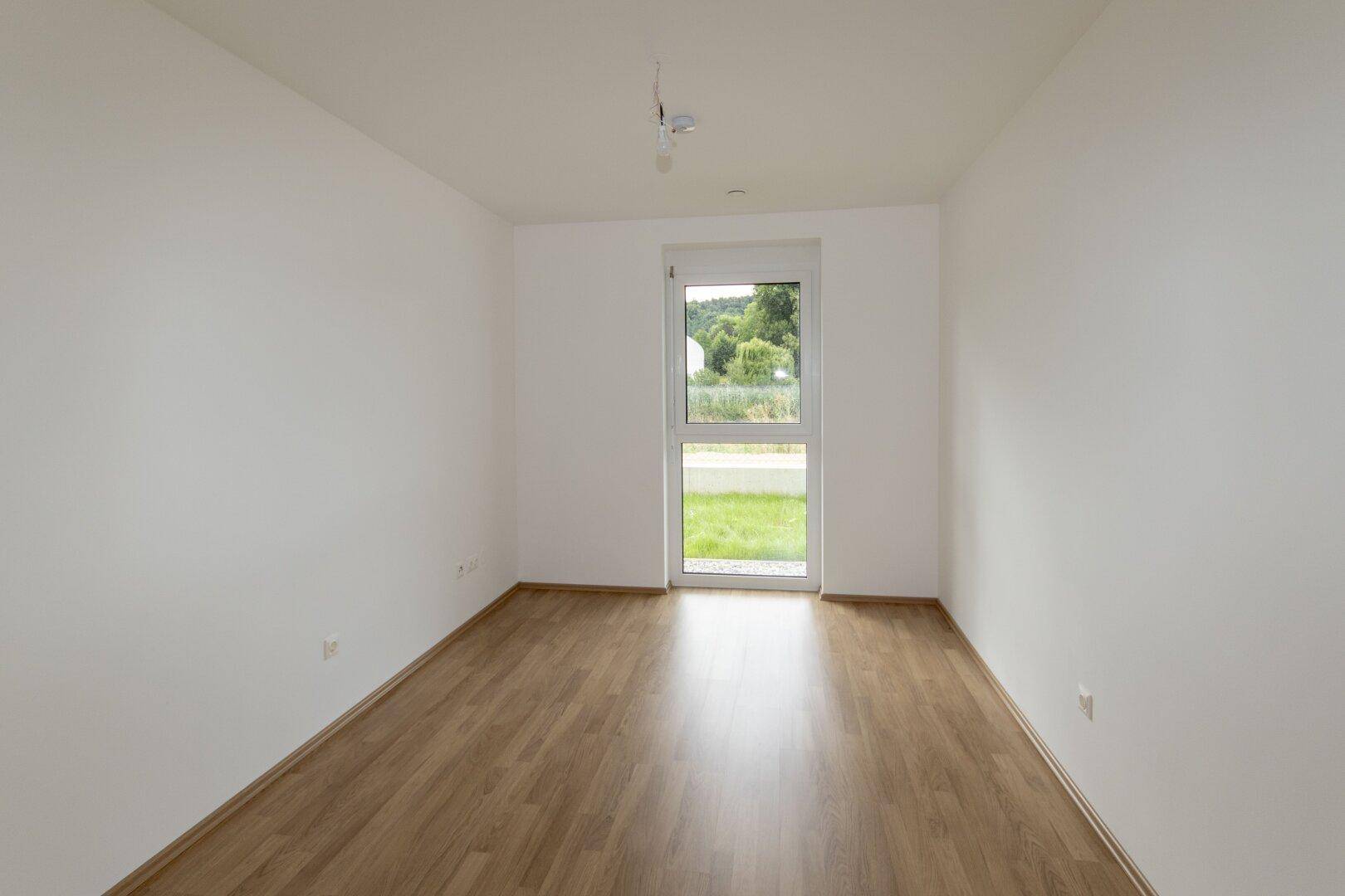 Helles Schlafzimmer mit Laminatboden und Fenster mit Blick ins Grüne.