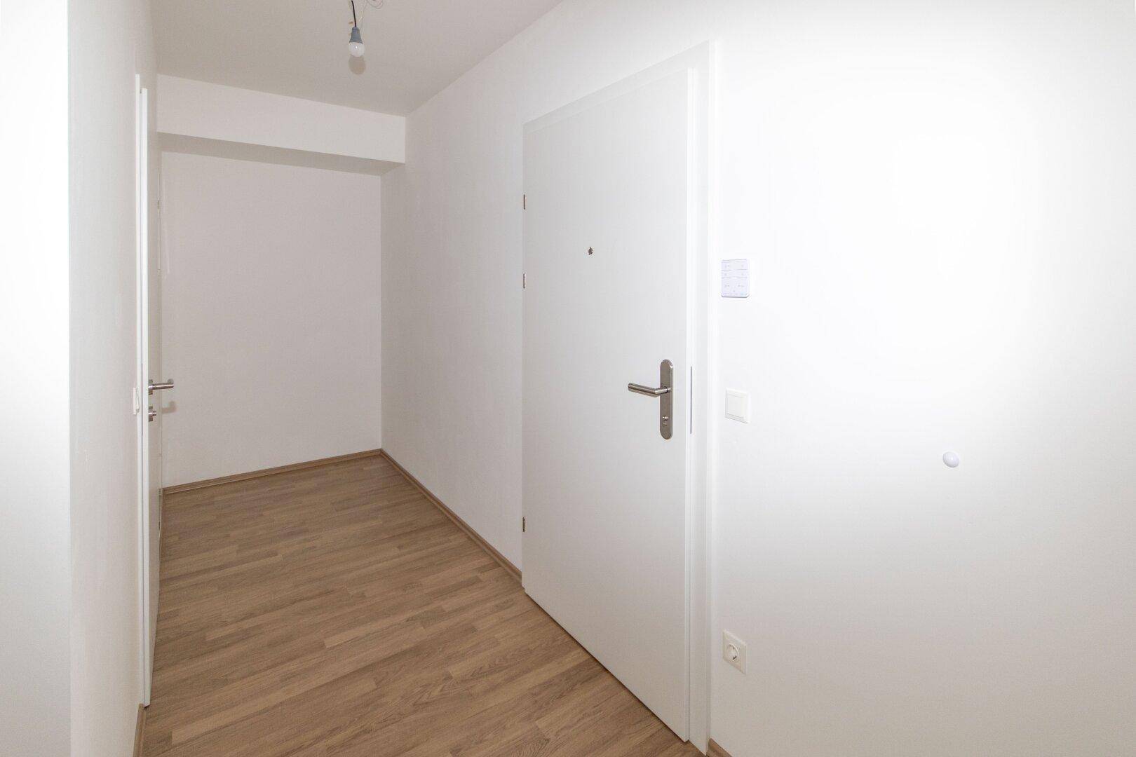 Heller Eingangsbereich mit weißer Tür und modernem Laminatboden, der zu weiteren Räumen führt.