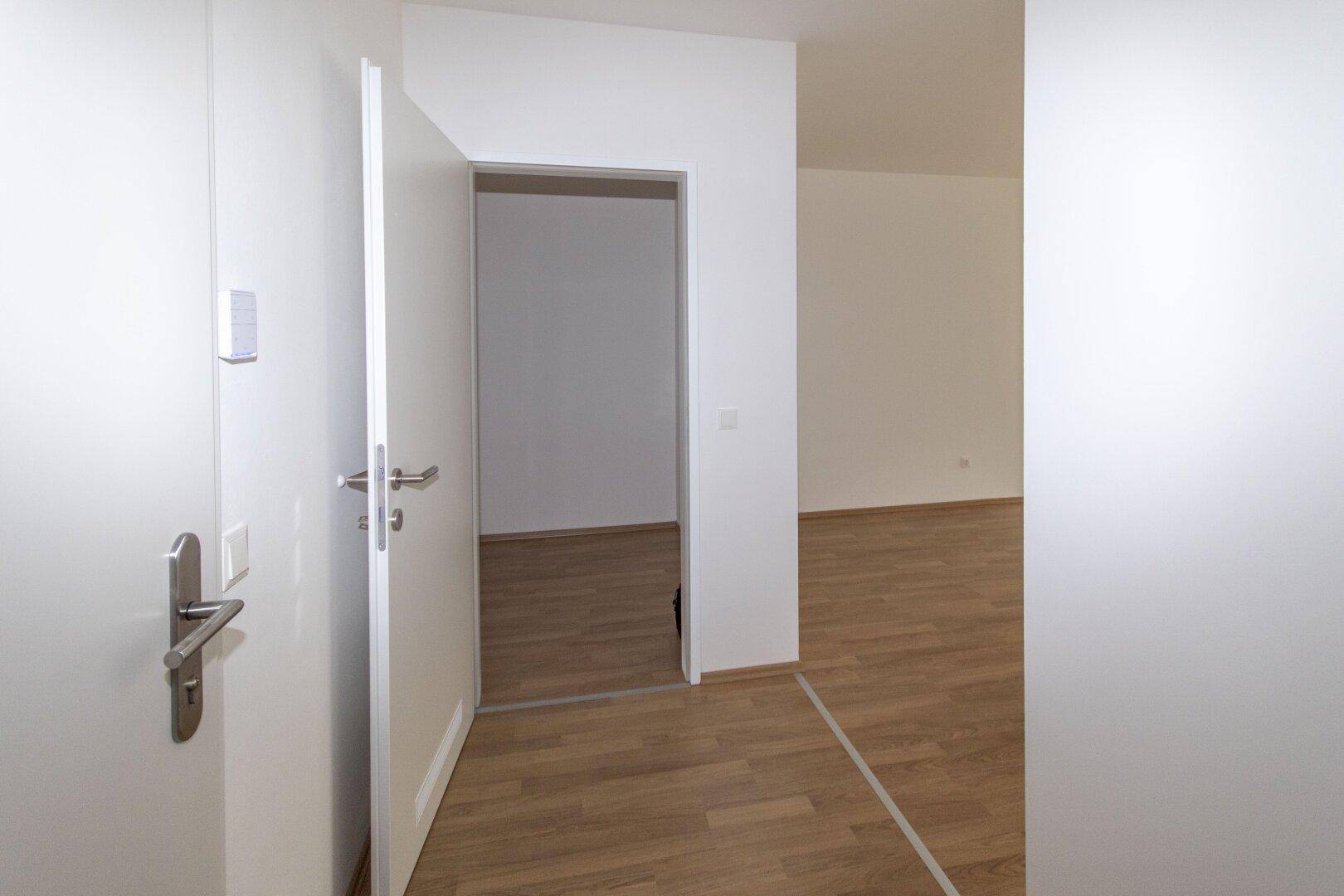 Blick aus einem hellen Flur in einen angrenzenden Raum mit identischem Laminatboden und weißen Wänden.