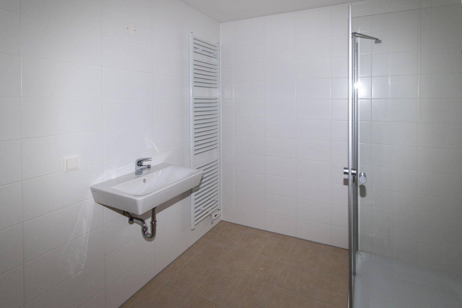 Funktionales Badezimmer mit Waschbecken, beheiztem Handtuchhalter und Duschbereich, hell gefliest.