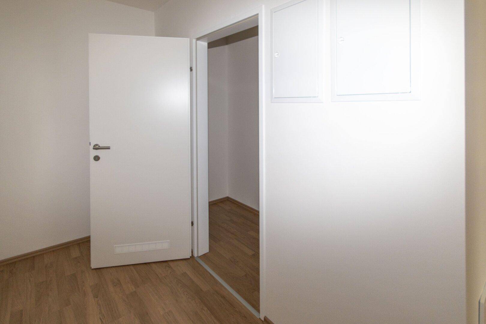Schlichter Flur mit Laminatboden und weiß gestrichenen Wänden, der in einen weiteren Raum führt.