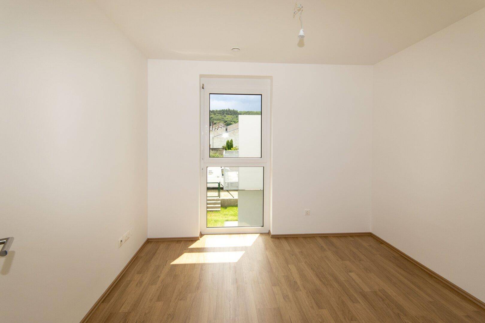 Helles Schlafzimmer mit Laminatboden und großem Fenster, das viel Tageslicht hereinlässt.