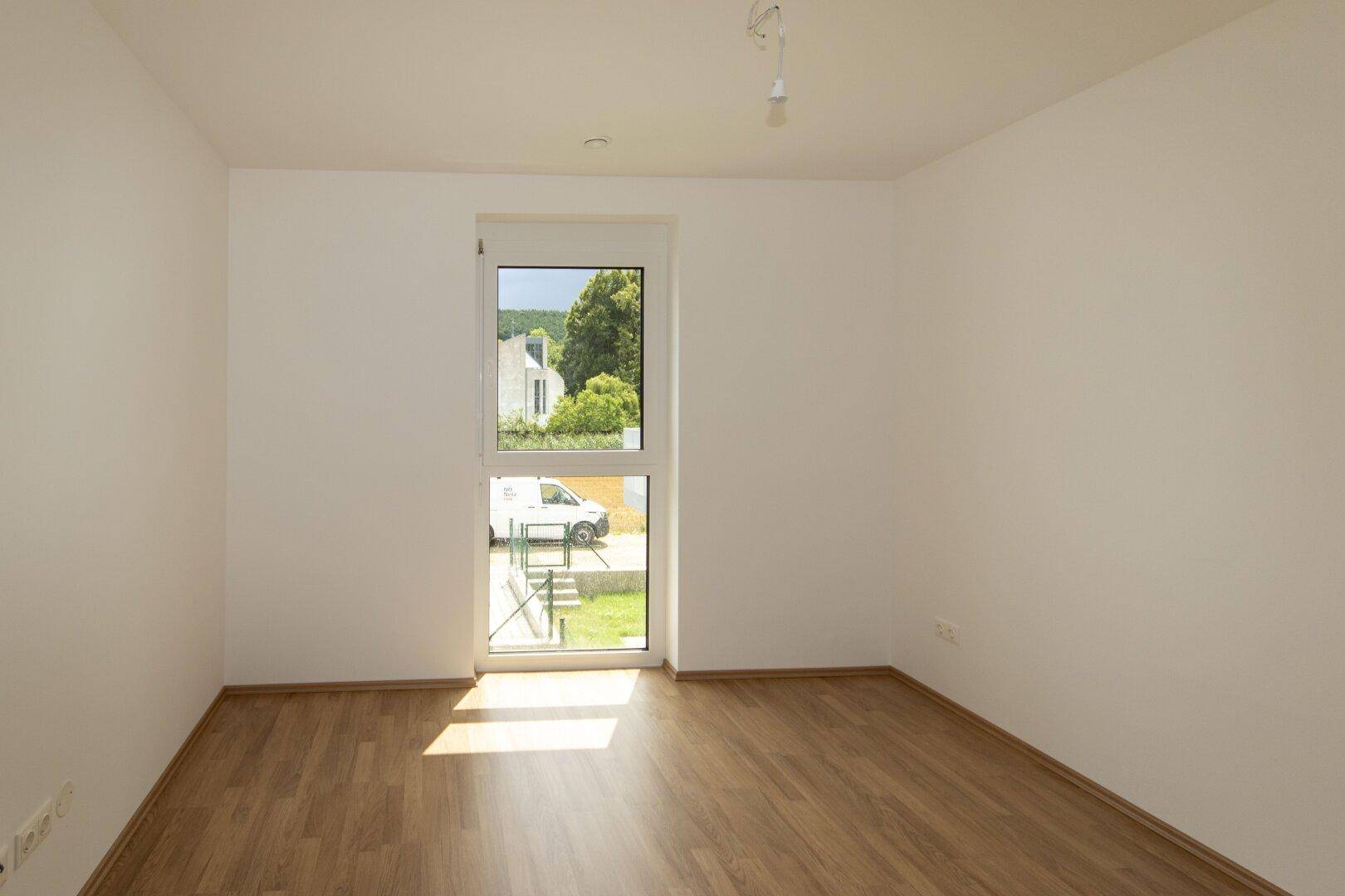Unmöbliertes Zimmer mit Laminatboden und Fenster, das einen Blick ins Grüne bietet.