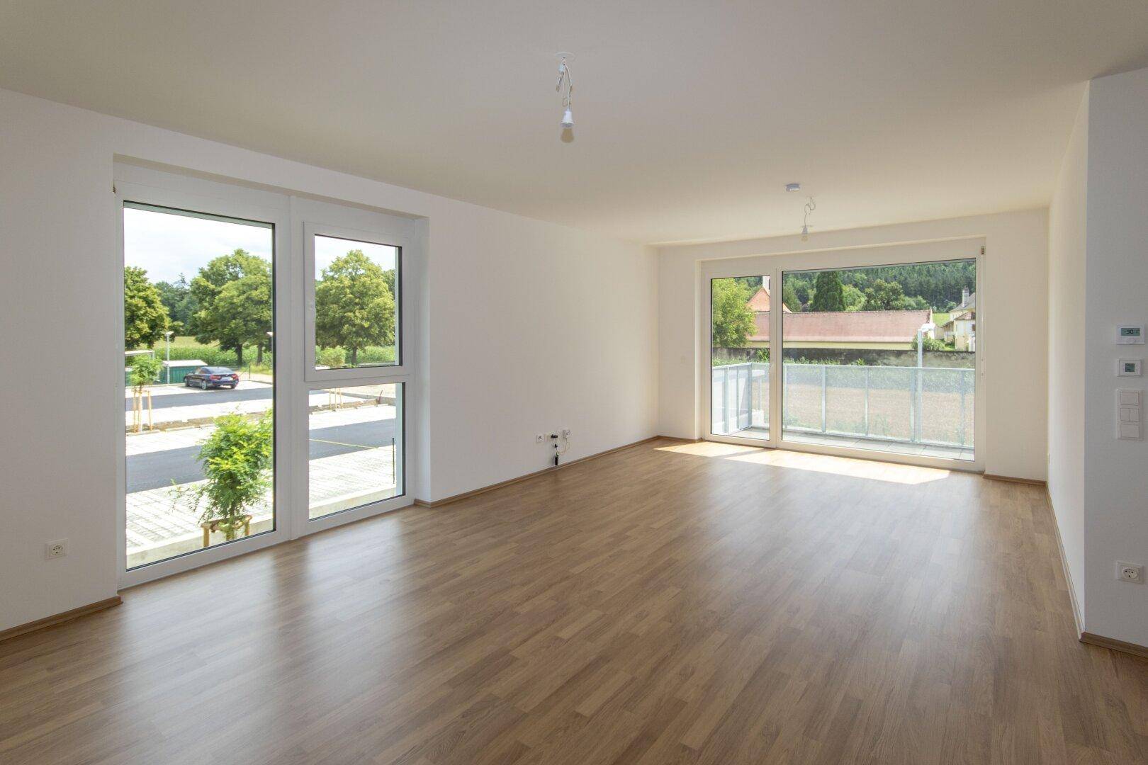 Helles, leeres Wohnzimmer mit großen Fenstern und modernem Laminatboden.
