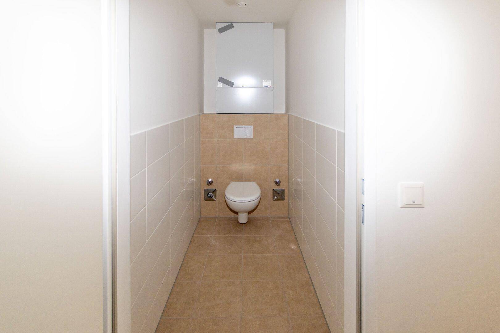 Separates WC mit wandhängender Toilette und hellen Fliesen an Wänden und Boden.
