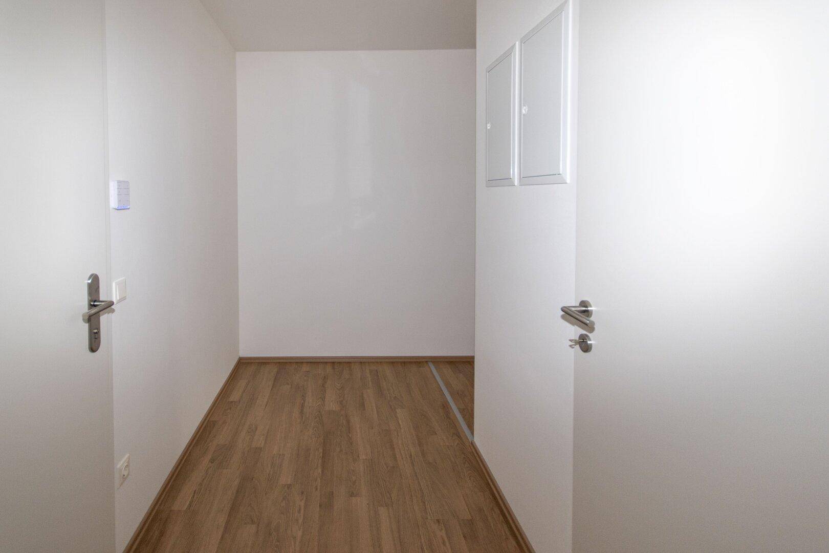 Schmaler Flur mit Laminatboden und weißen Wänden, der zu weiteren Räumen führt.