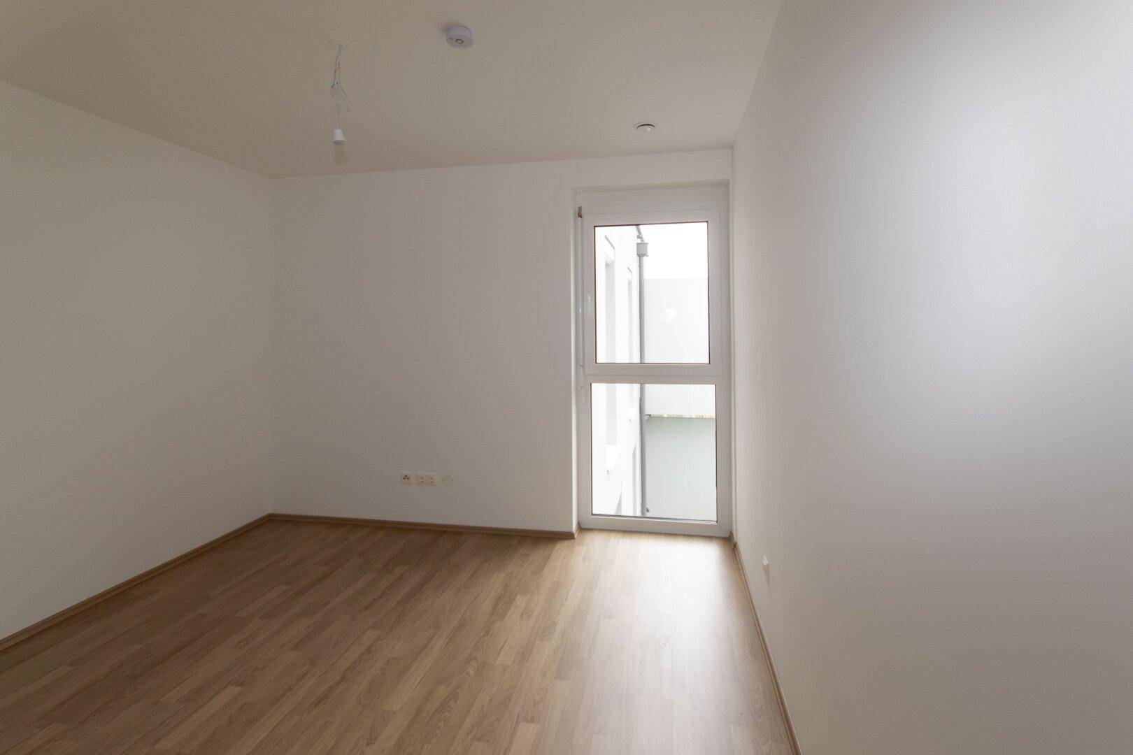 Helles, leeres Schlafzimmer mit Laminatboden und einem hohen Fenster, das viel Tageslicht spendet.