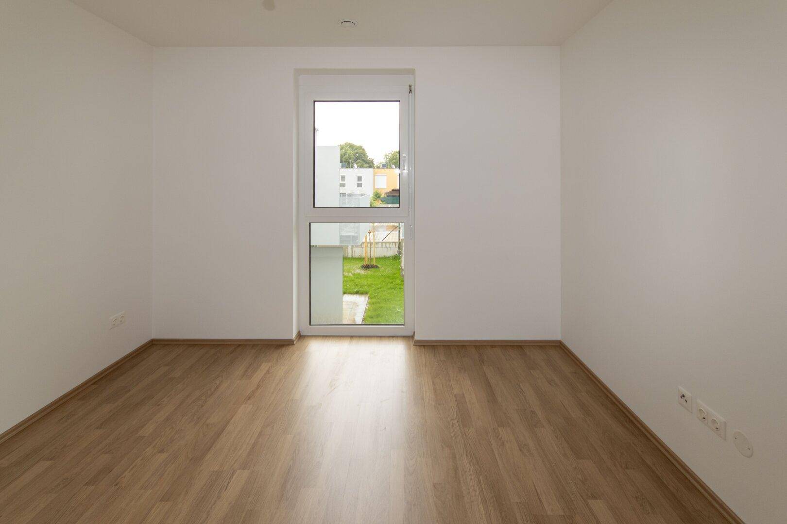 Unmöbliertes Schlafzimmer mit Laminatboden und Fenster, das einen Blick auf eine kleine Grünfläche bietet.