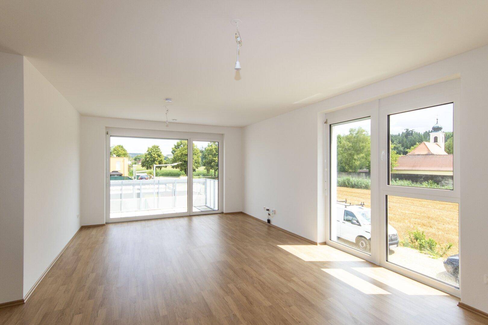 Helles, leeres Wohnzimmer mit Laminatboden und großen Fenstern, die einen weiten Blick ins Grüne bieten.