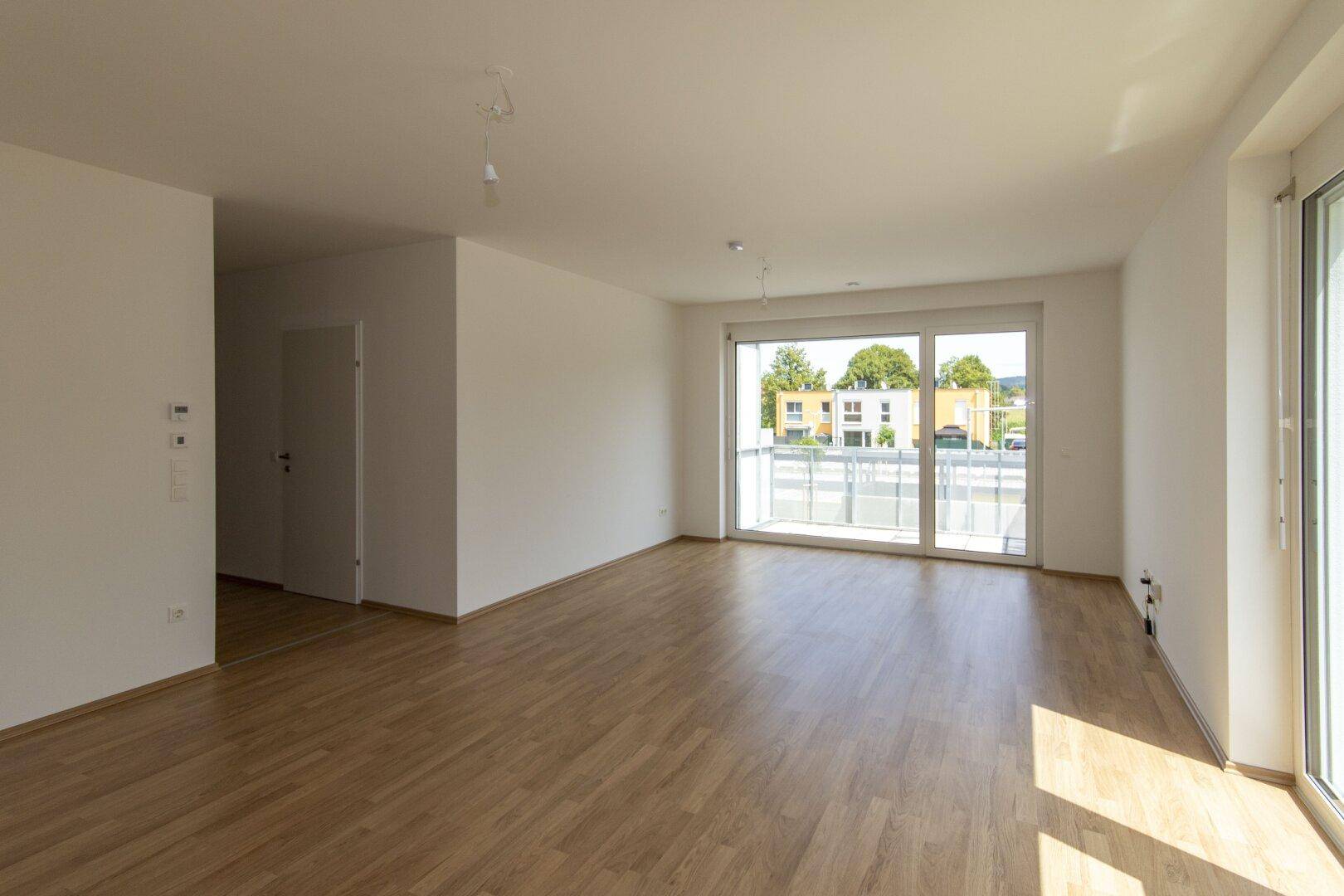 Geräumiges, leeres Wohnzimmer mit Laminatboden und großen Fenstern, die viel Tageslicht hereinlassen.