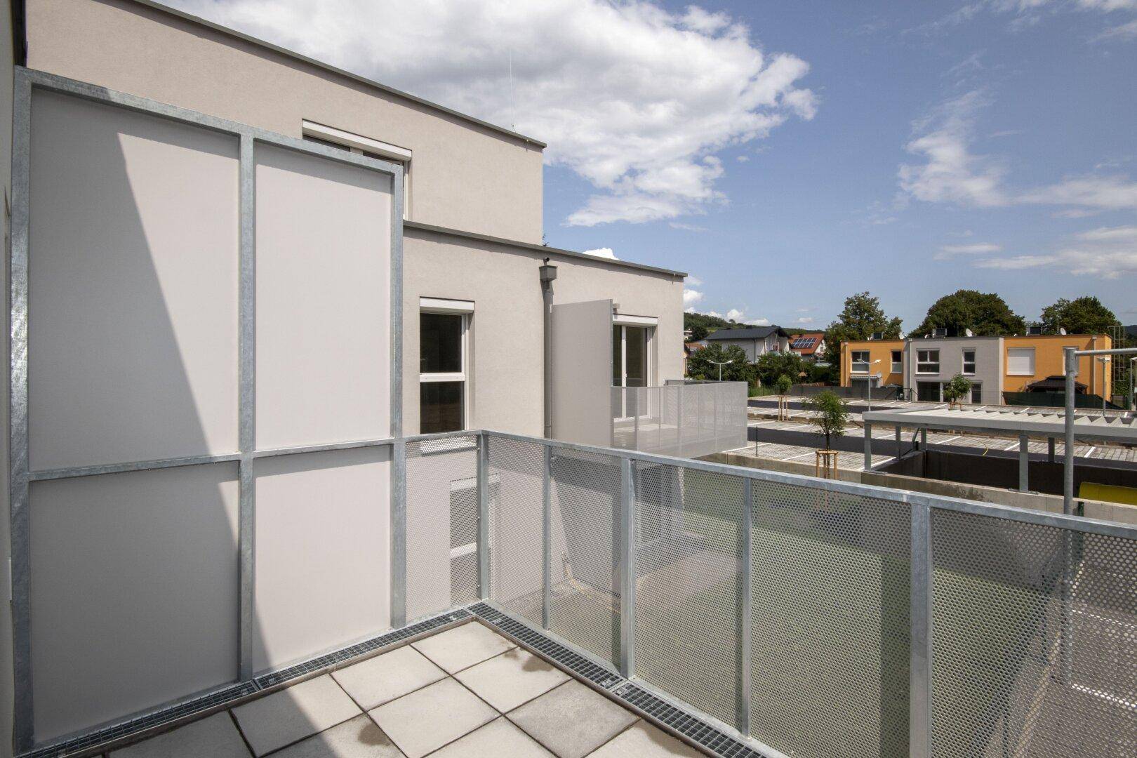 Terrasse mit Fliesenboden und Metallgeländer, mit Blick auf benachbarte Gebäude und einen Carport.