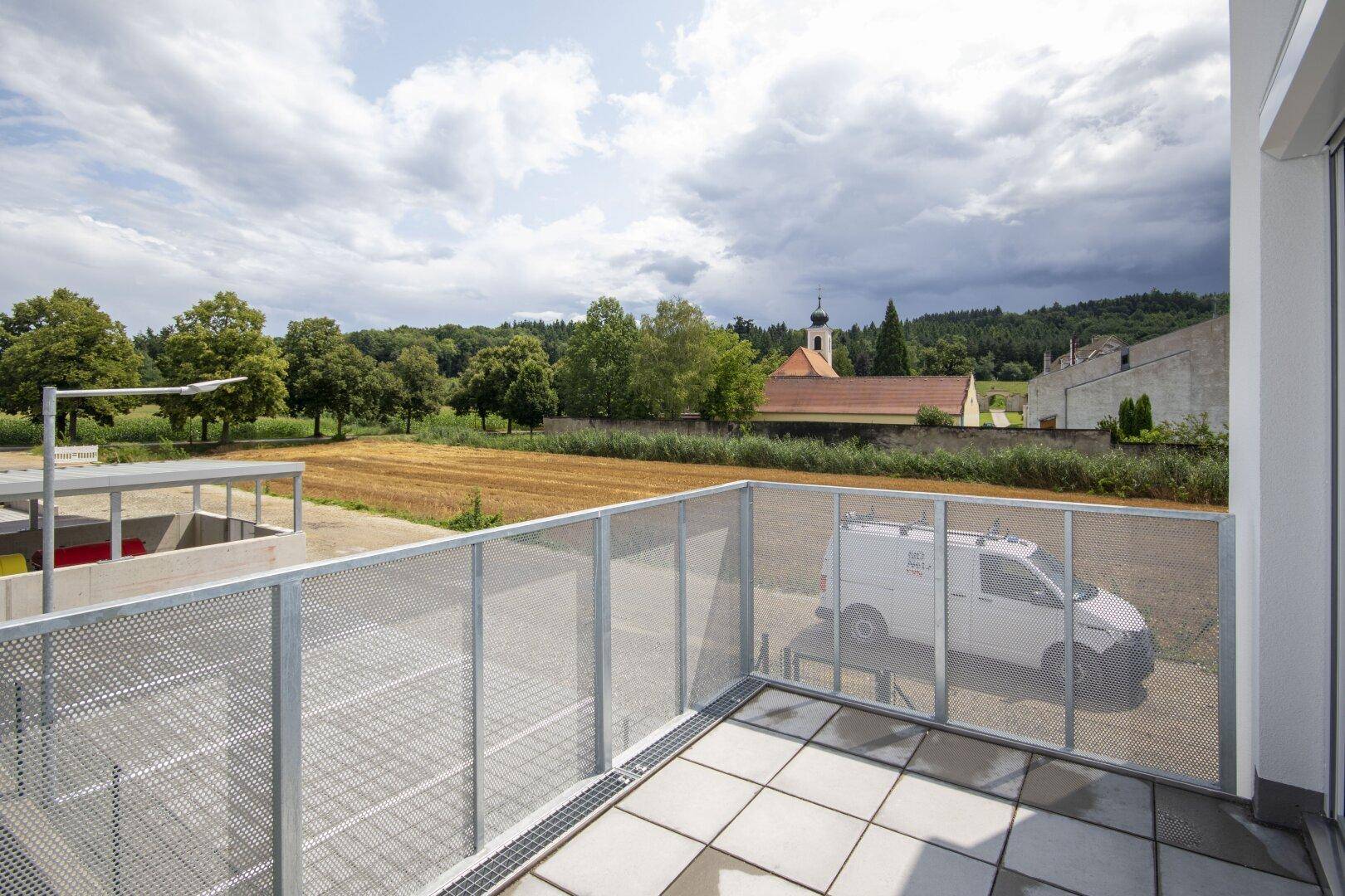Terrasse mit Fliesenboden und Metallgeländer, bietet einen weiten Blick über Felder und eine Kirche.