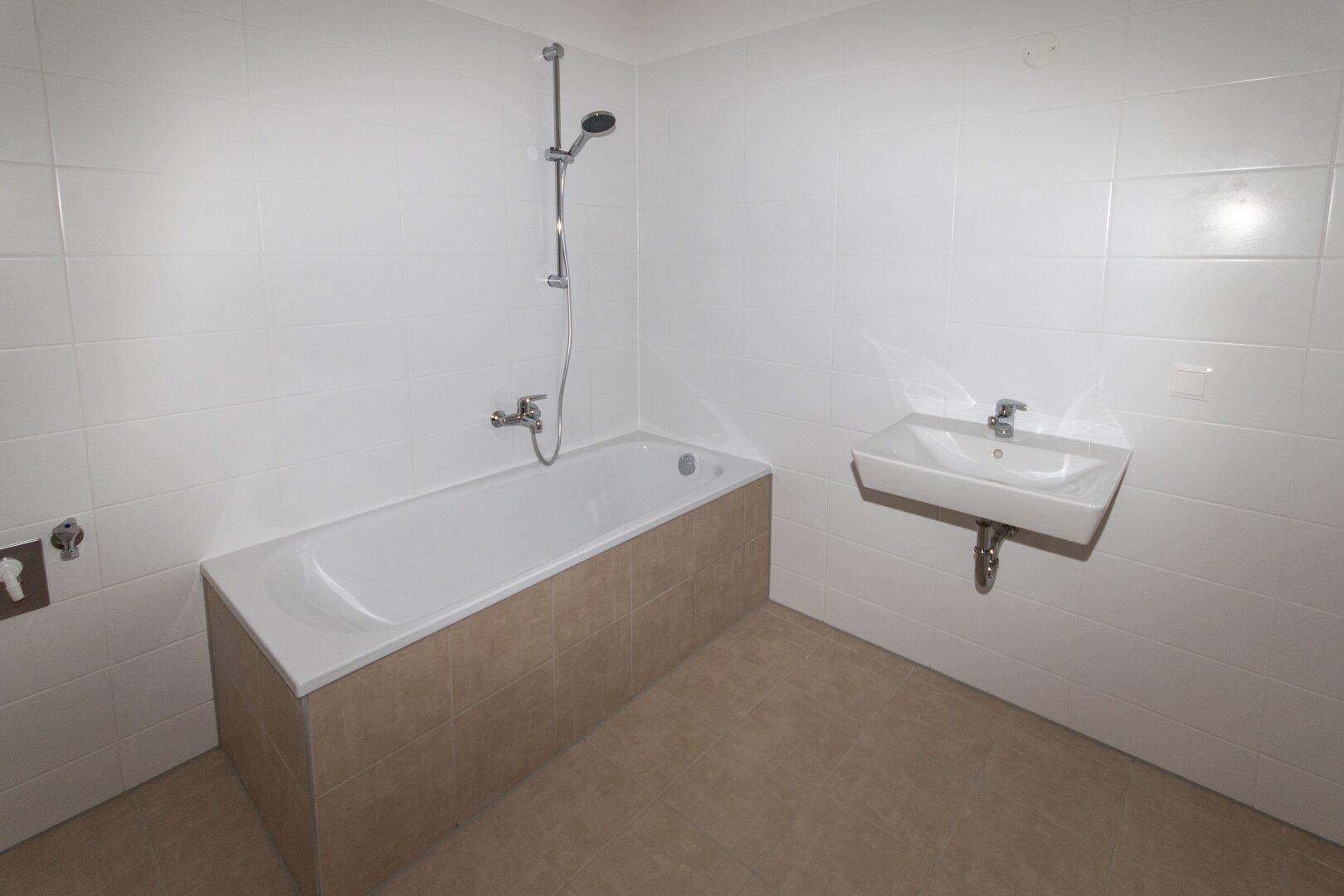 Modernes Badezimmer mit Badewanne, Duscharmatur und Einzelwaschbecken, komplett gefliest in Weiß und Beige.