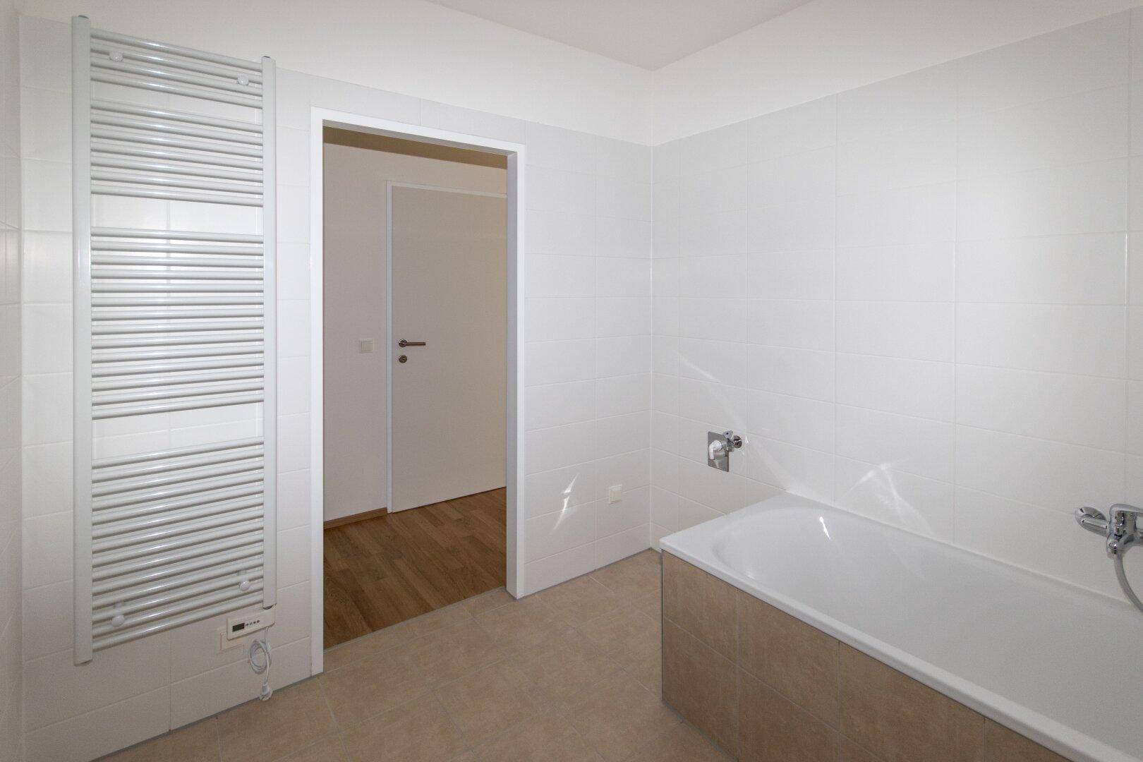 Badezimmer mit Badewanne und beheiztem Handtuchhalter, gefliest in Weiß und Beige, mit Zugang zu einem weiteren Raum.