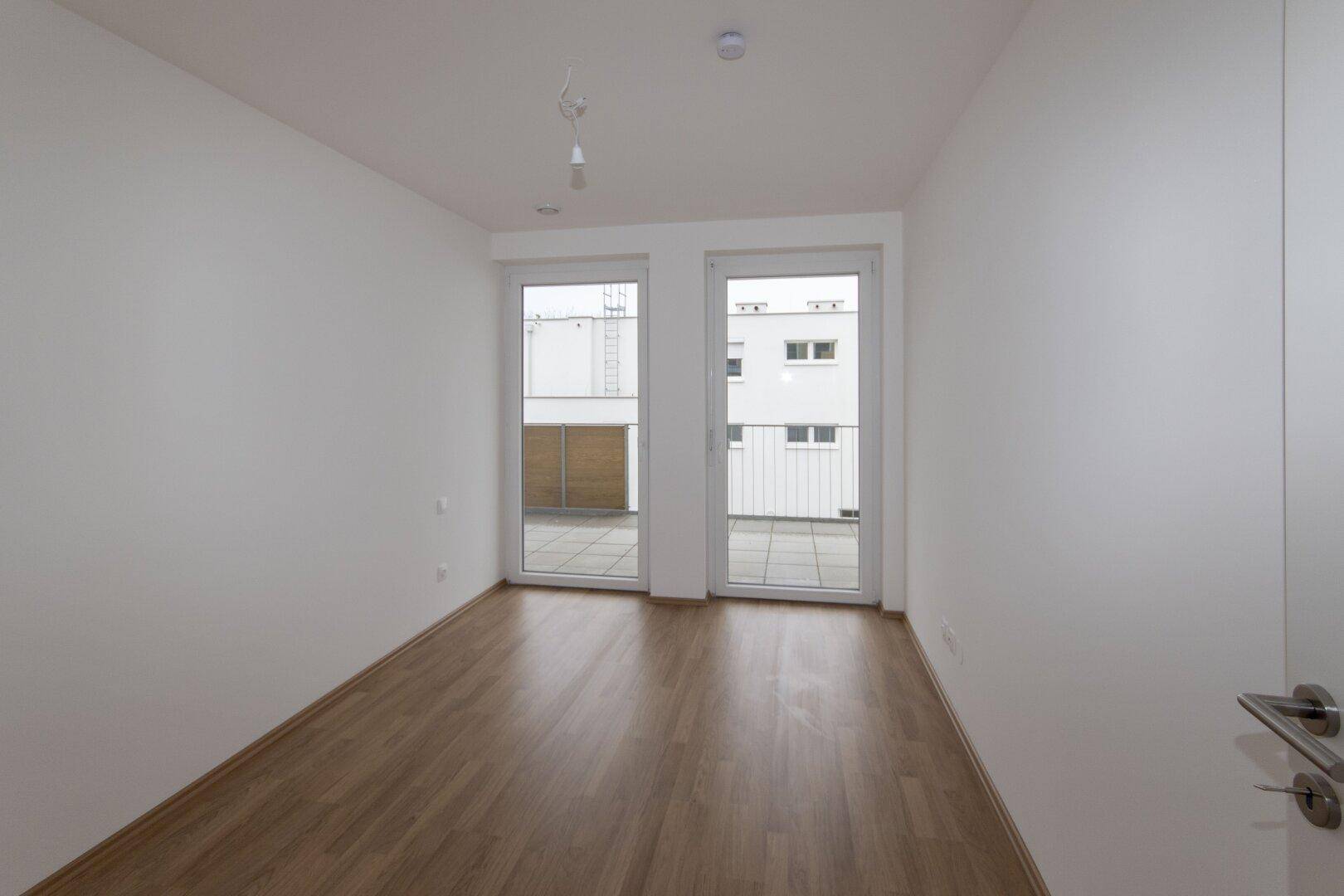 Helles, leeres Schlafzimmer mit Holzboden und großen Fenstern zum Balkon.