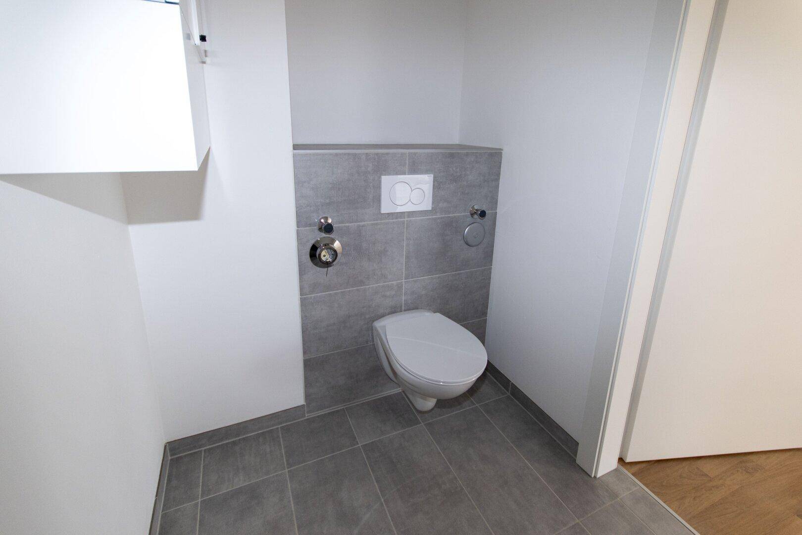 Separates WC mit Wand-WC und modernen grauen Fliesen, sauber und funktional.
