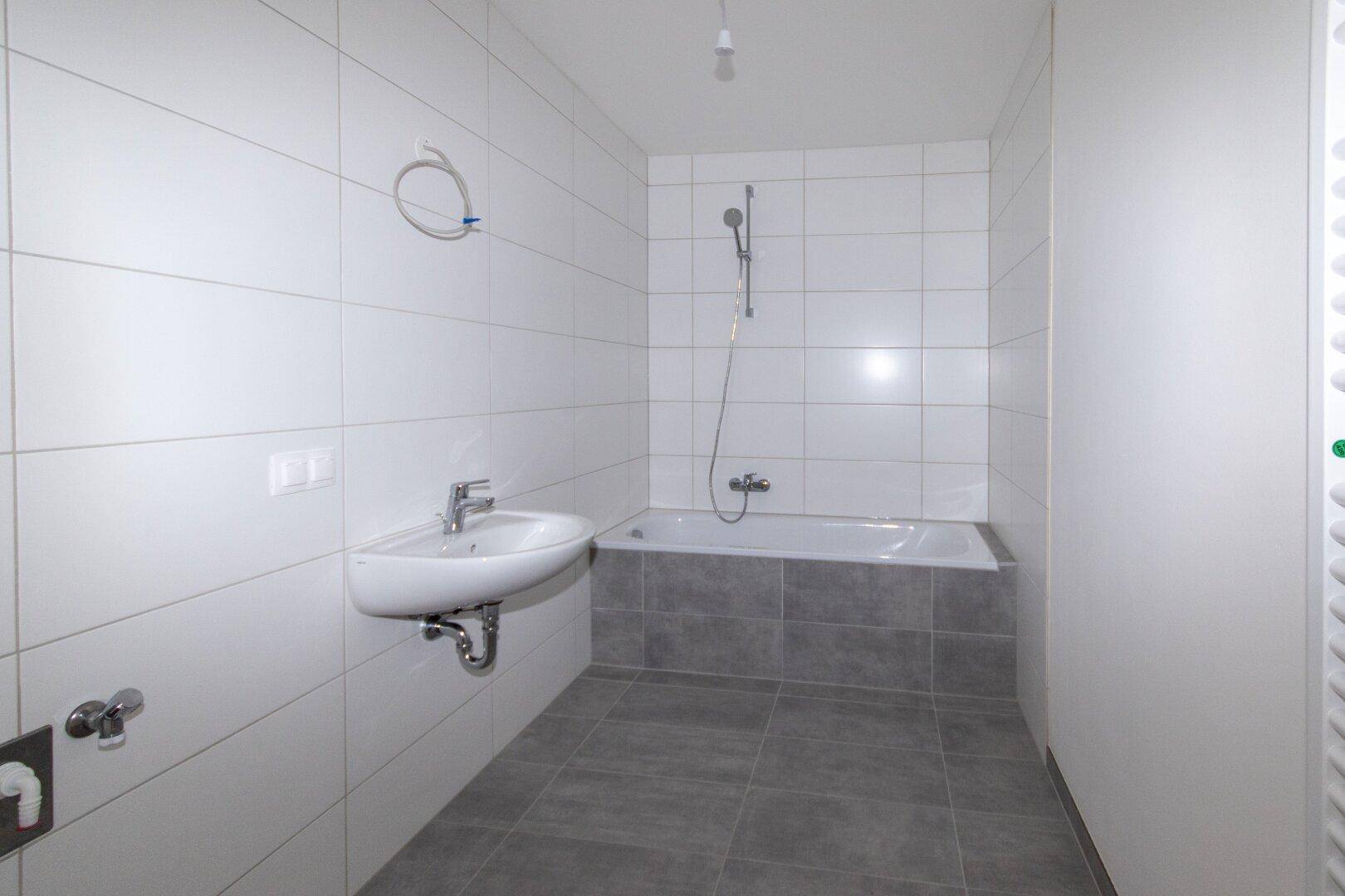 Modernes Badezimmer mit weißen Wandfliesen, grauen Bodenfliesen und einer Badewanne.