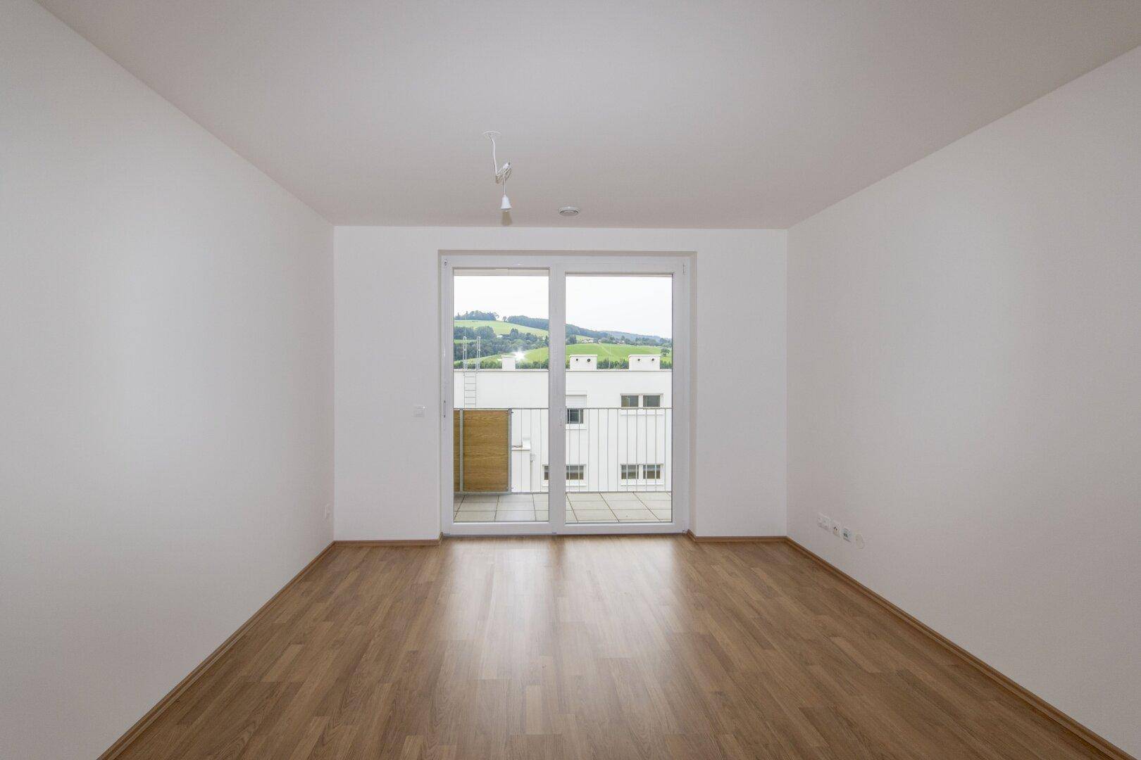 Heller Wohnraum mit Laminatboden und großen Schiebetüren, die Zugang zu einem Balkon mit grüner Aussicht bieten.
