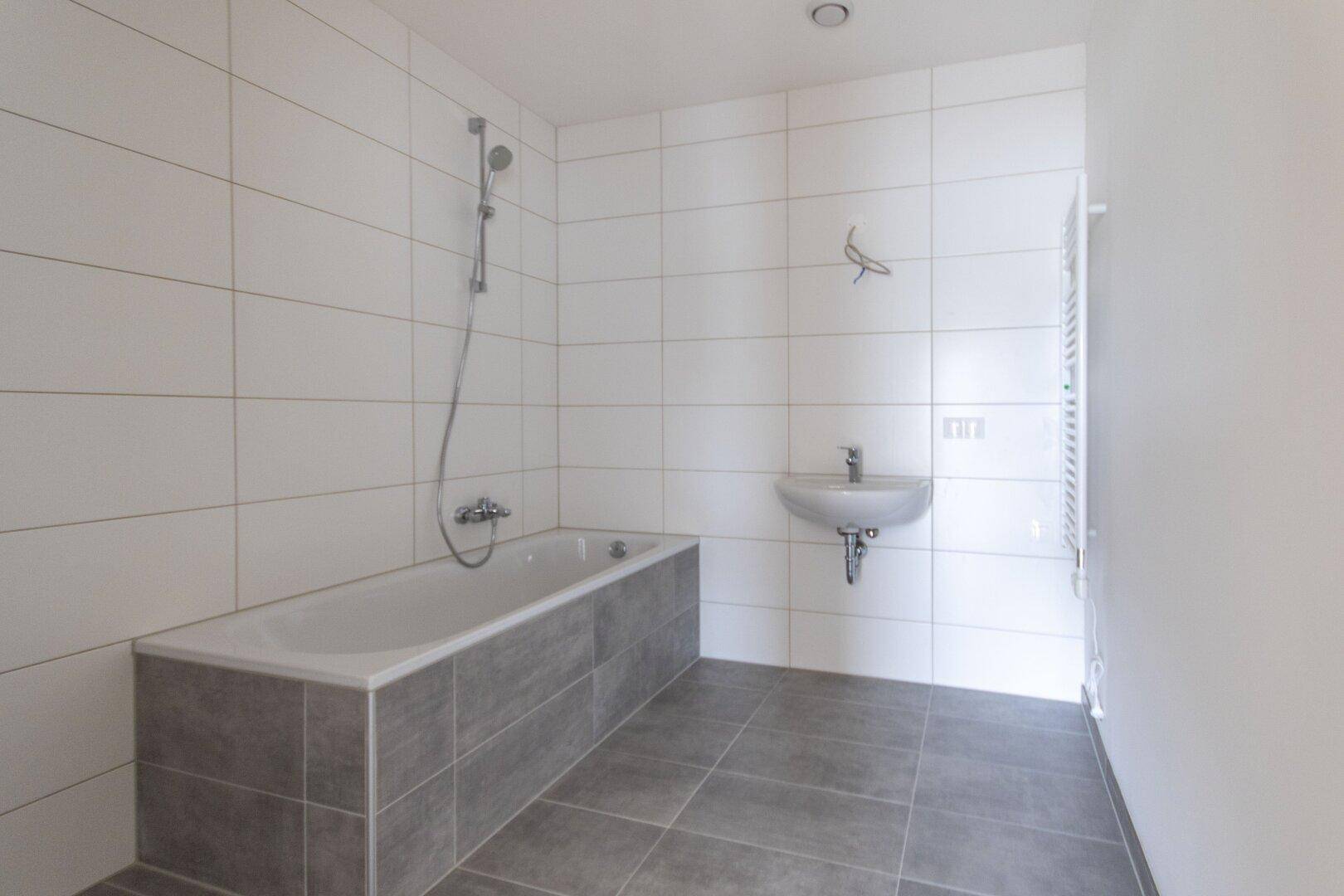 Modernes Badezimmer mit weißen Wandfliesen, grauen Bodenfliesen, einer Badewanne mit Duschfunktion und einem Waschbecken.