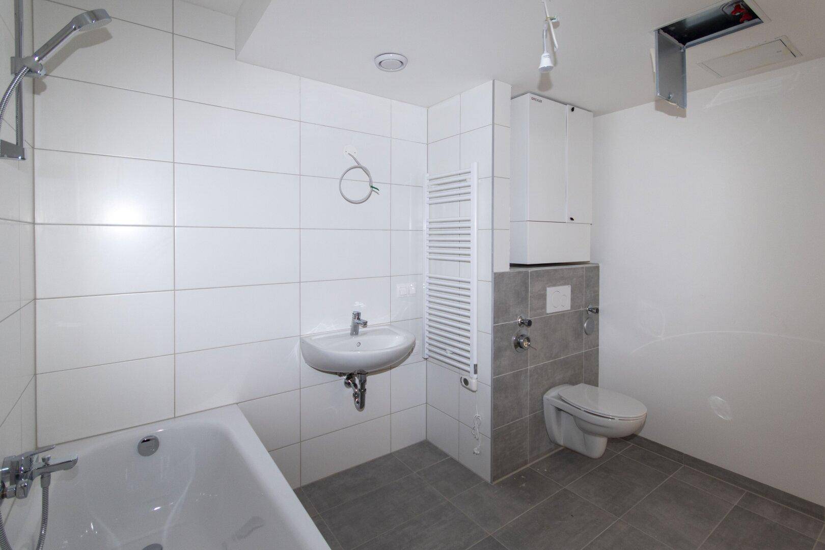 Modernes Badezimmer mit Badewanne, Duscharmatur, WC und Handtuchheizkörper in Weiß und Grau gefliest.