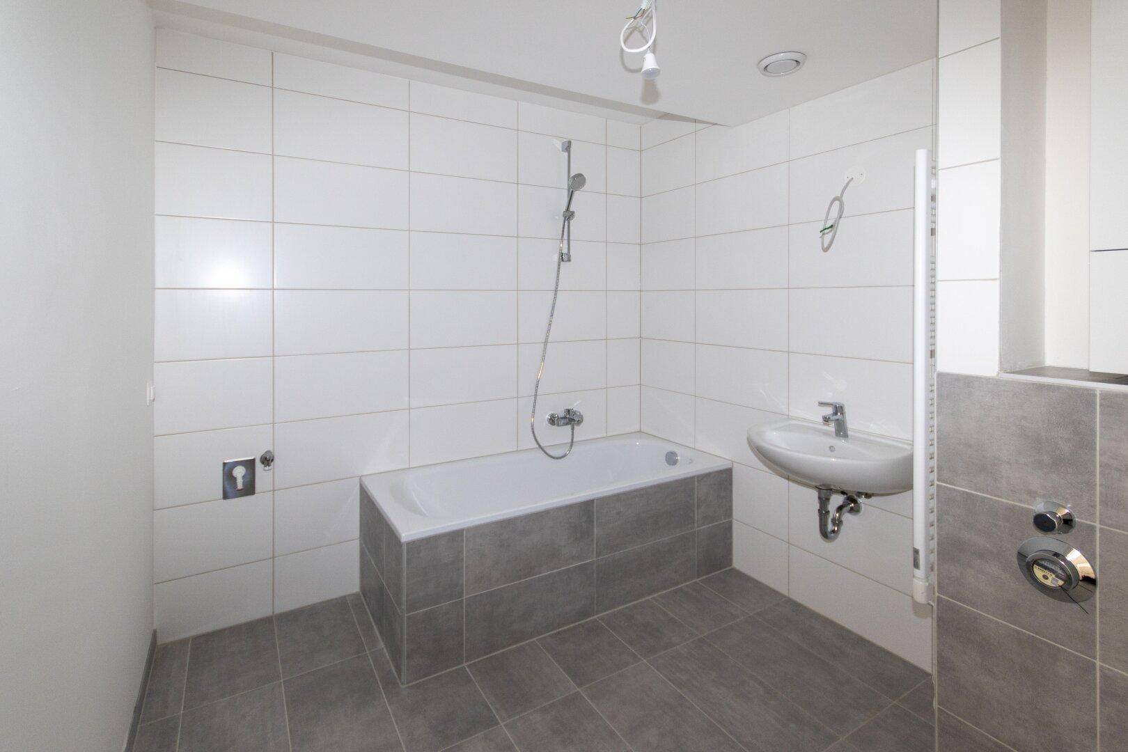 Helles Badezimmer mit Badewanne, Duscharmatur und modernen Fliesen in Weiß und Grau, unbenutzt.