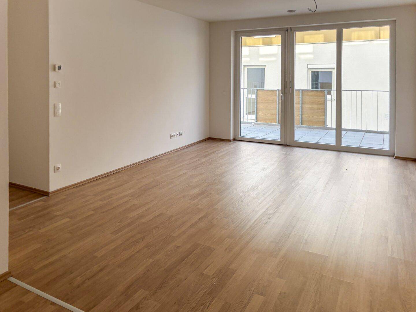 Weitläufiger, heller Raum mit Laminatboden und breiten Terrassentüren, ideal für einen Wohnbereich.
