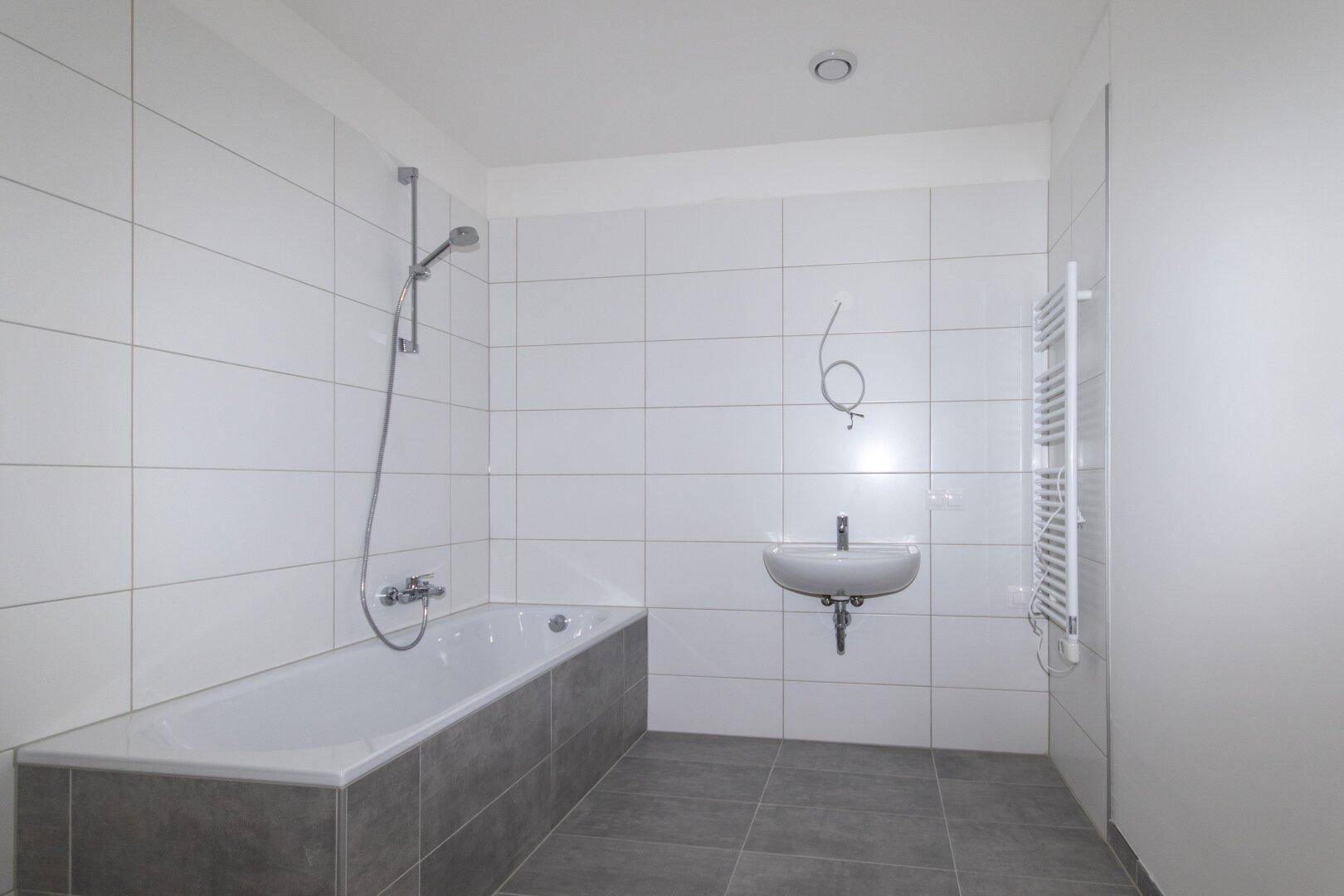 Modernes Badezimmer mit weißen Wandfliesen, grauen Bodenfliesen und einer Badewanne mit Duschfunktion.