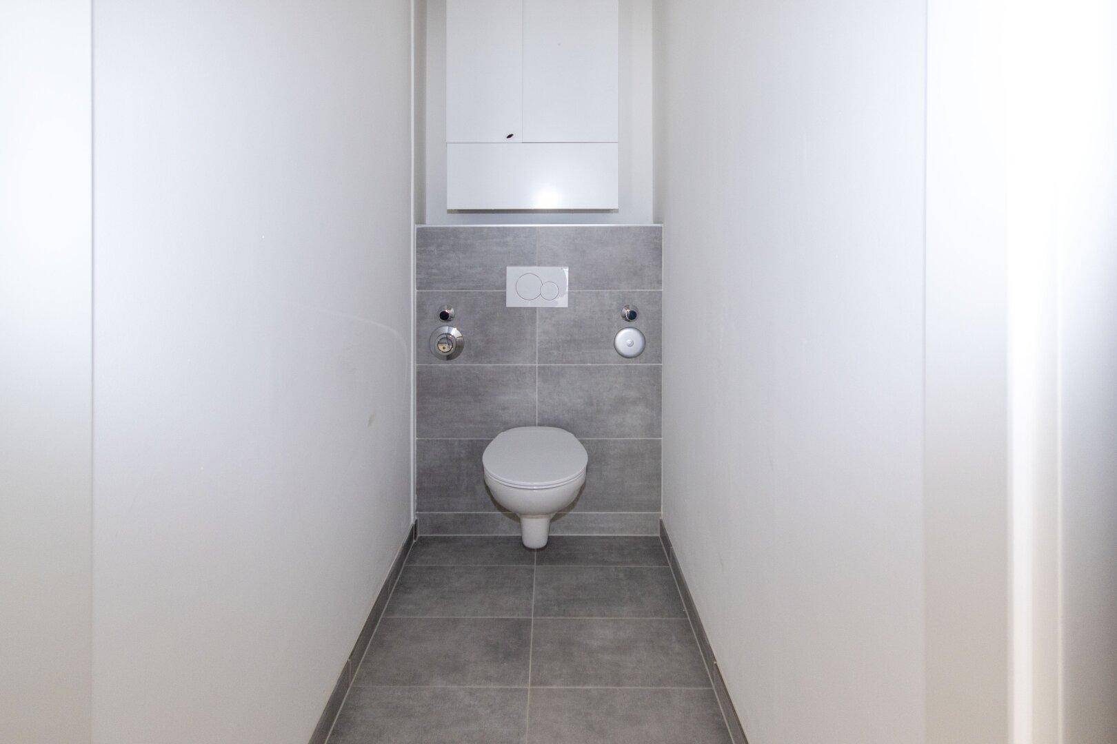 Separates WC mit wandhängender Toilette, grauen Fliesen und einem kleinen Wandschrank.
