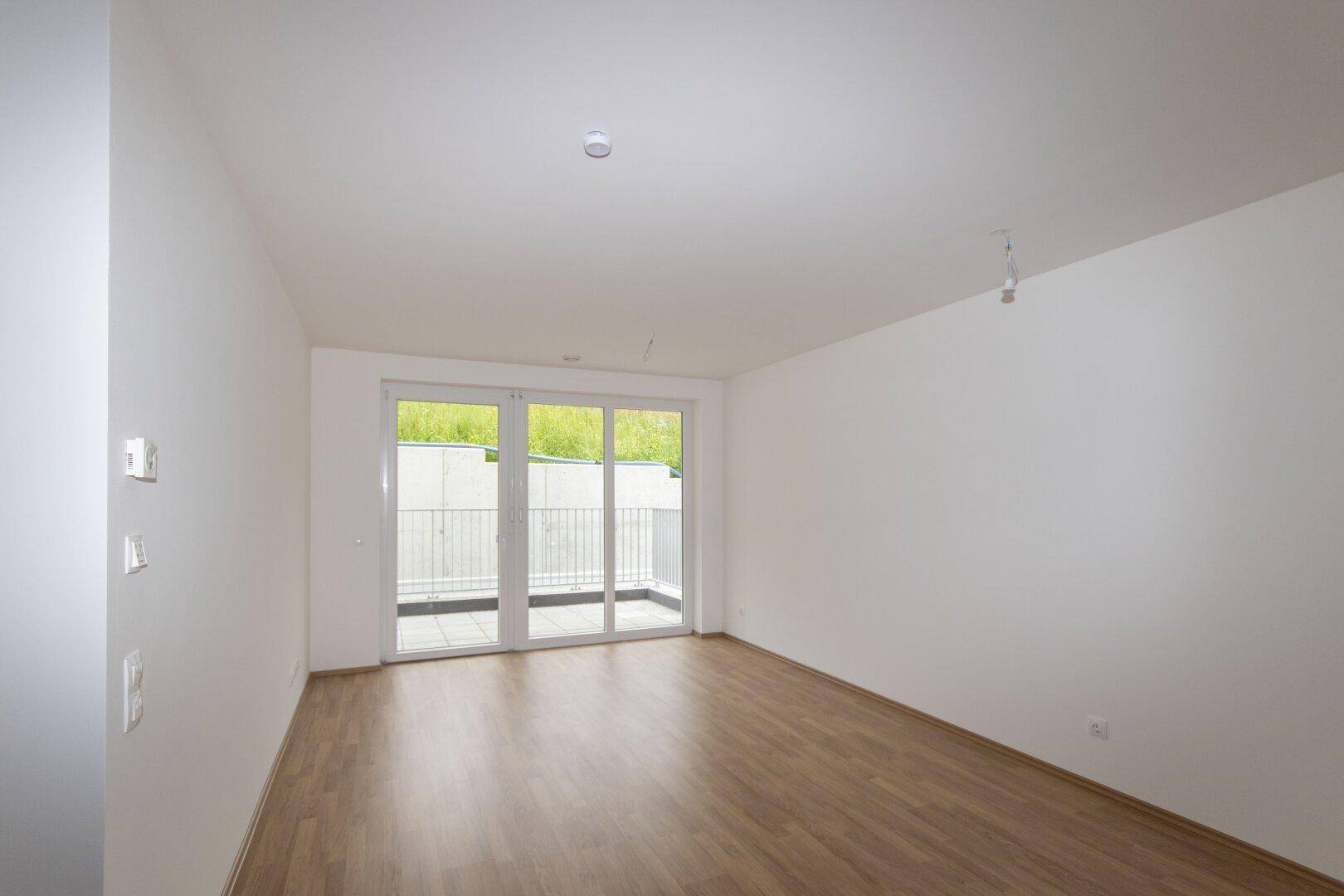 Helles, leeres Zimmer mit Laminatboden und großem Fenster mit Zugang zu einem Balkon.