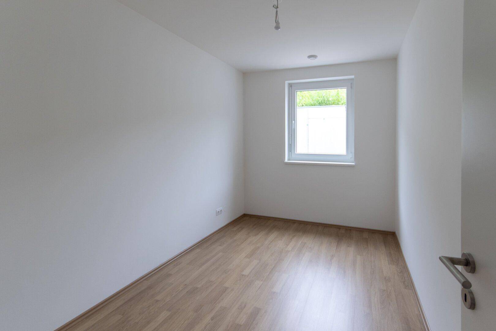 Unmöbliertes Zimmer mit Laminatboden, einem Fenster und weißen Wänden, ideal als Schlafzimmer.
