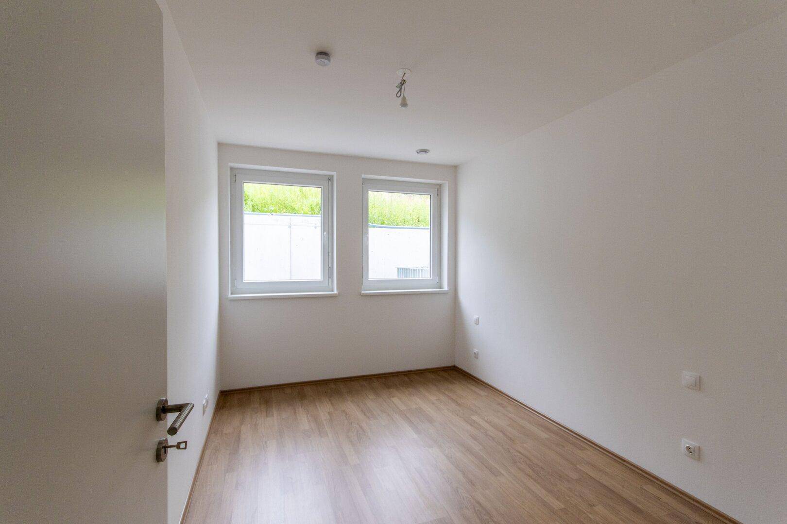 Helles Schlafzimmer mit zwei Fenstern, Laminatboden und weißen Wänden, unmöbliert.