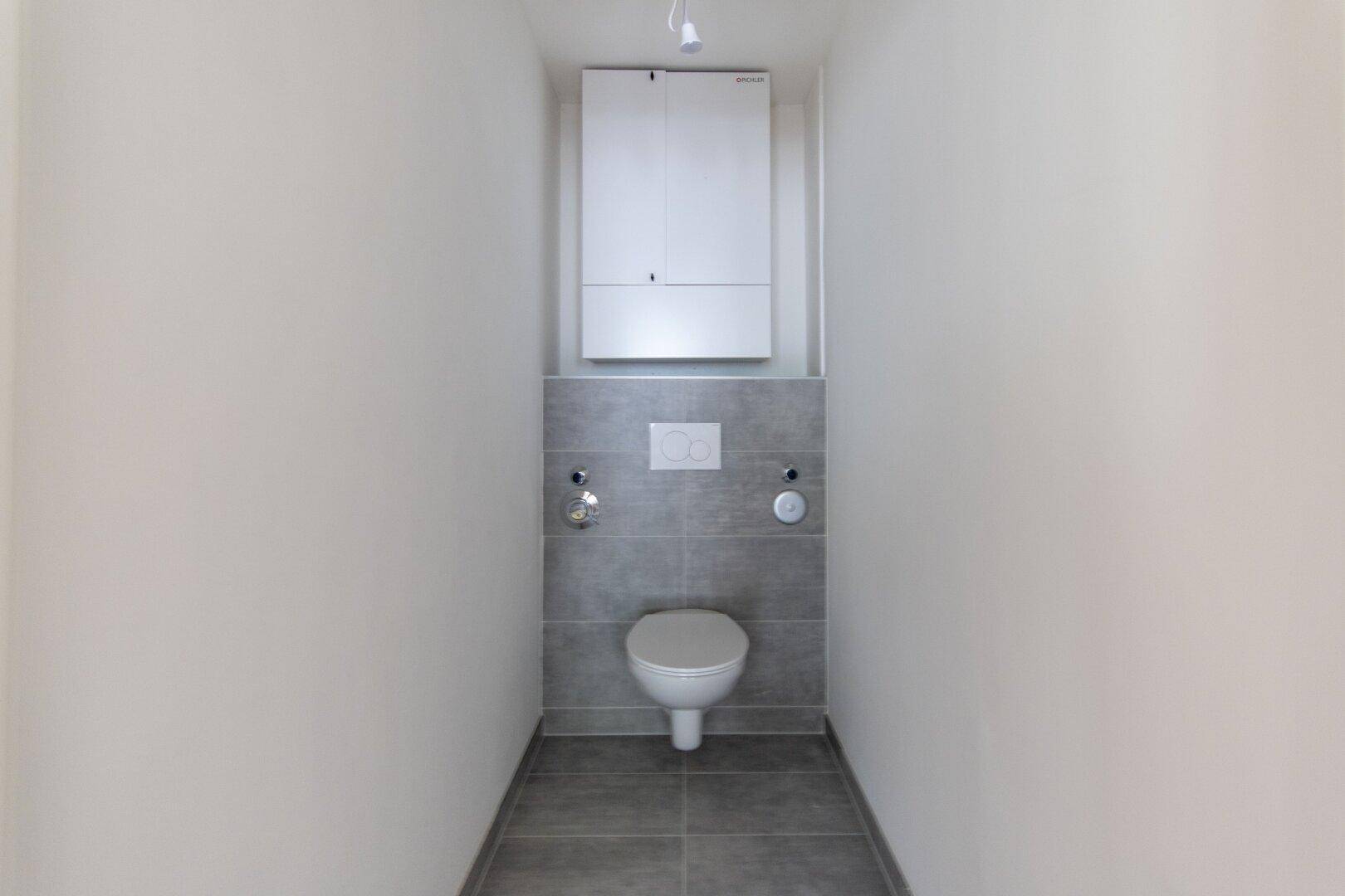 Separates WC mit wandhängender Toilette und grauen Fliesen im unteren Bereich.