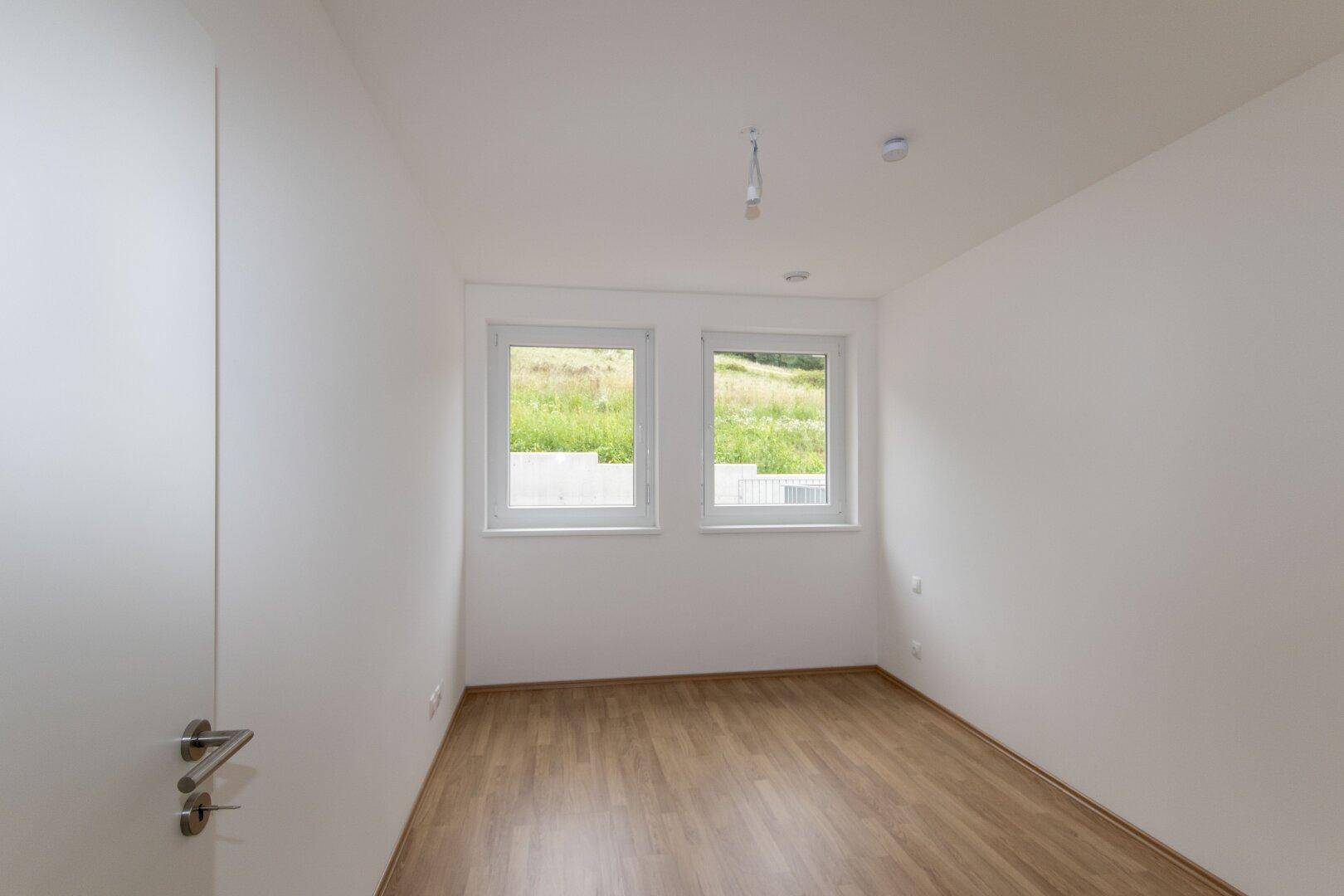 Helles Schlafzimmer mit Laminatboden, weißen Wänden und zwei Fenstern mit Blick ins Grüne.