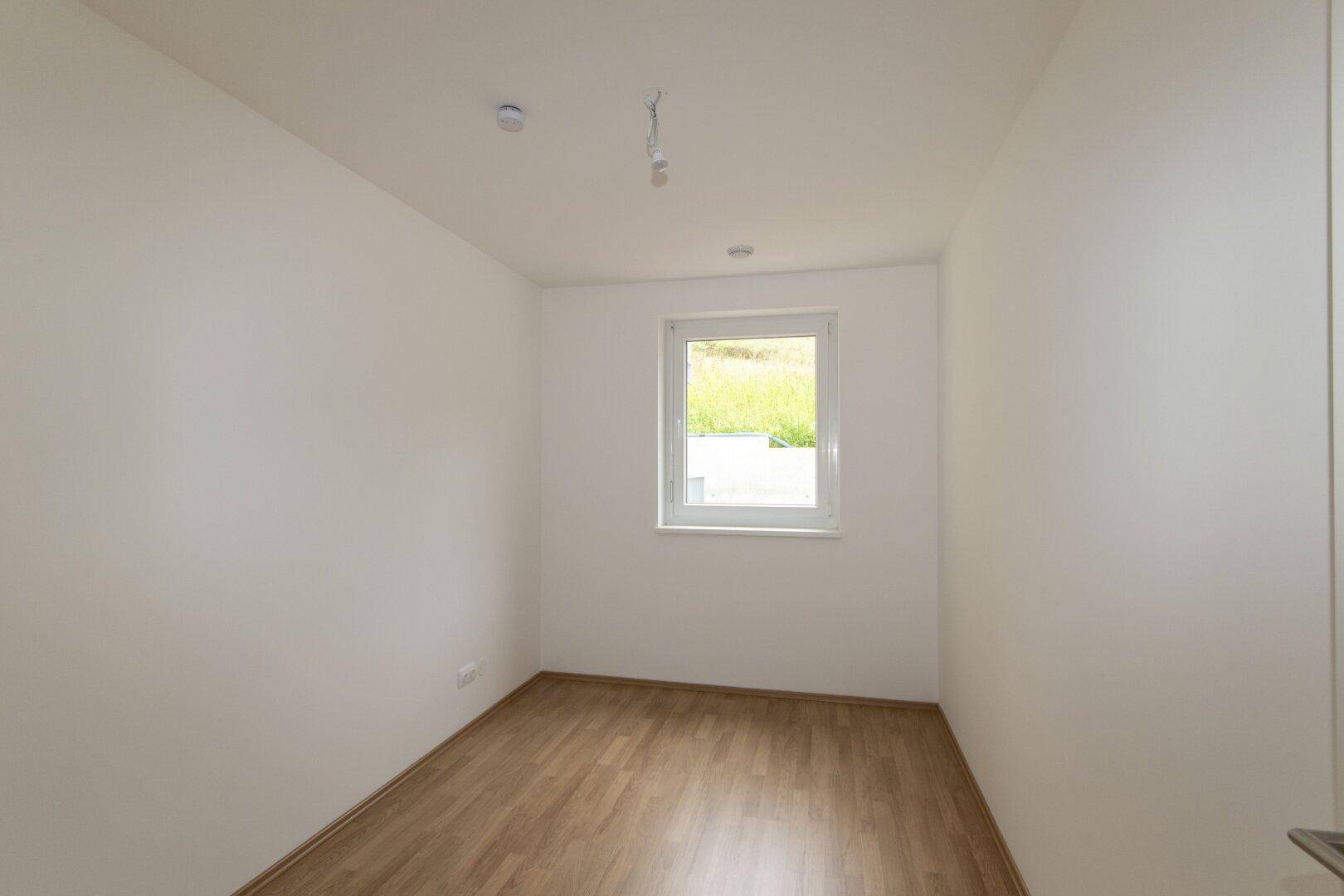Ruhiges Schlafzimmer mit Laminatboden, weißen Wänden und einem Fenster mit Blick auf die Natur.
