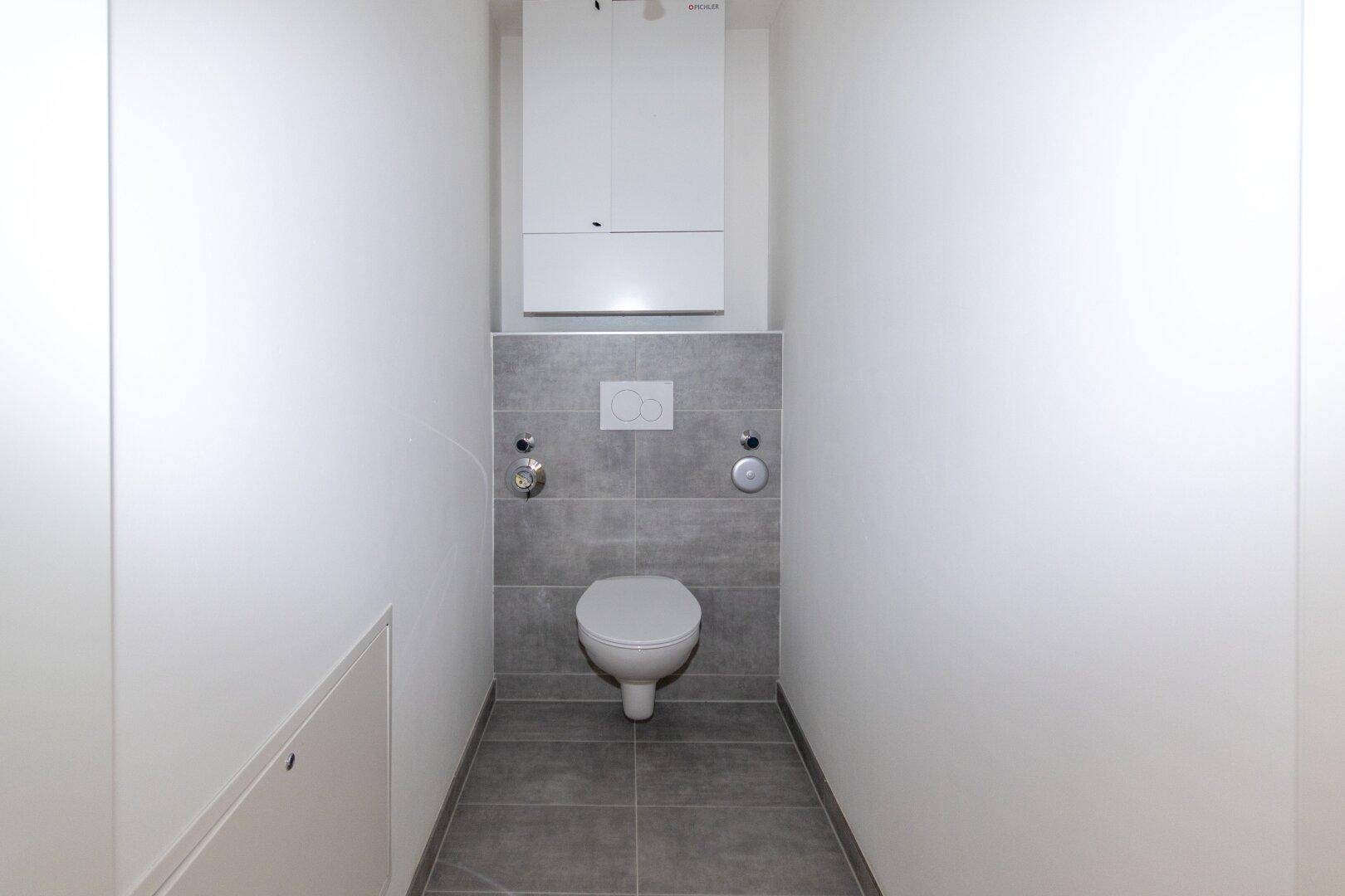 Separates WC mit wandhängender Toilette, grauen Fliesen und einem praktischen Wandschrank.
