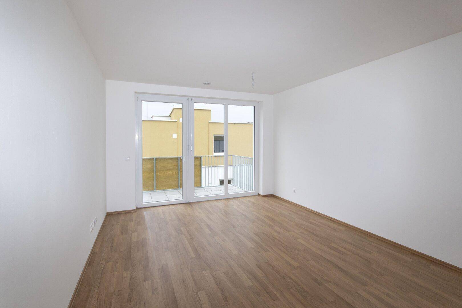 Heller, leerer Raum mit Holzboden und großem Fenster mit Zugang zum Balkon.