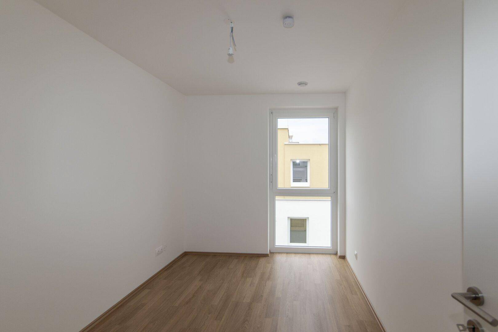 Helles Schlafzimmer mit Laminatboden und einem großen Fenster, das viel Tageslicht hereinlässt.