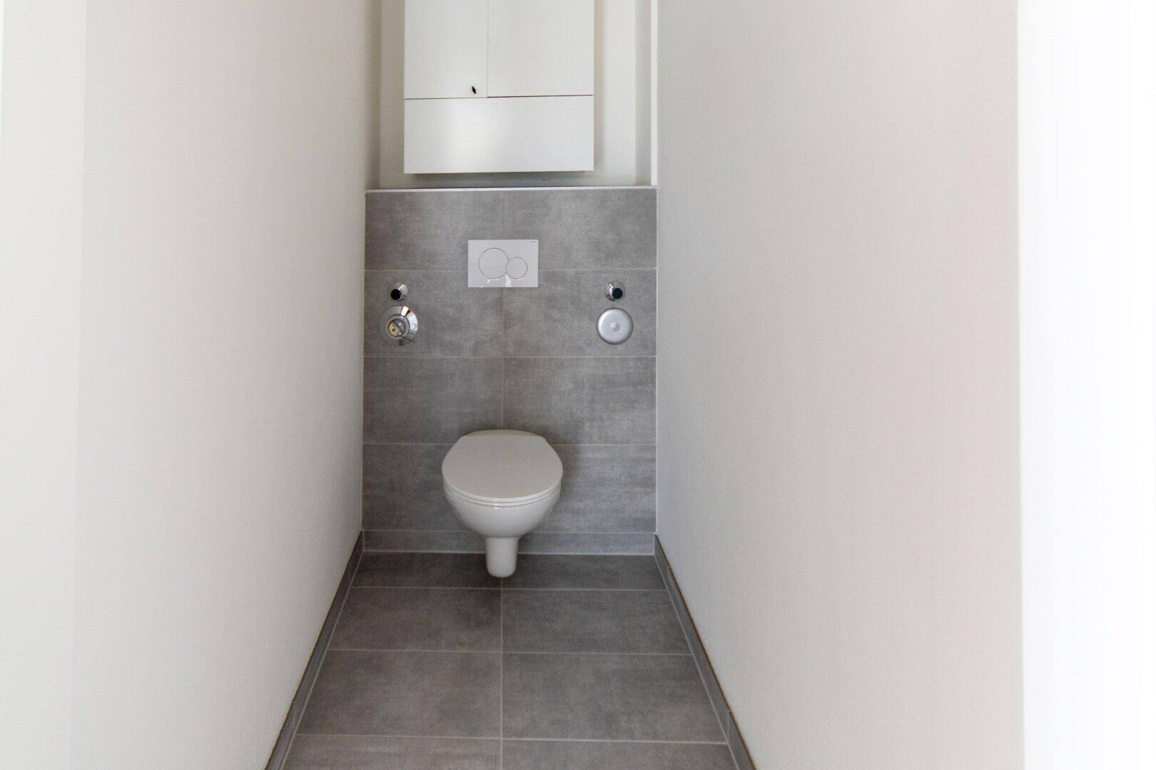 Separates WC mit wandhängender Toilette und grauen Fliesen, in einem modernen und gepflegten Zustand.