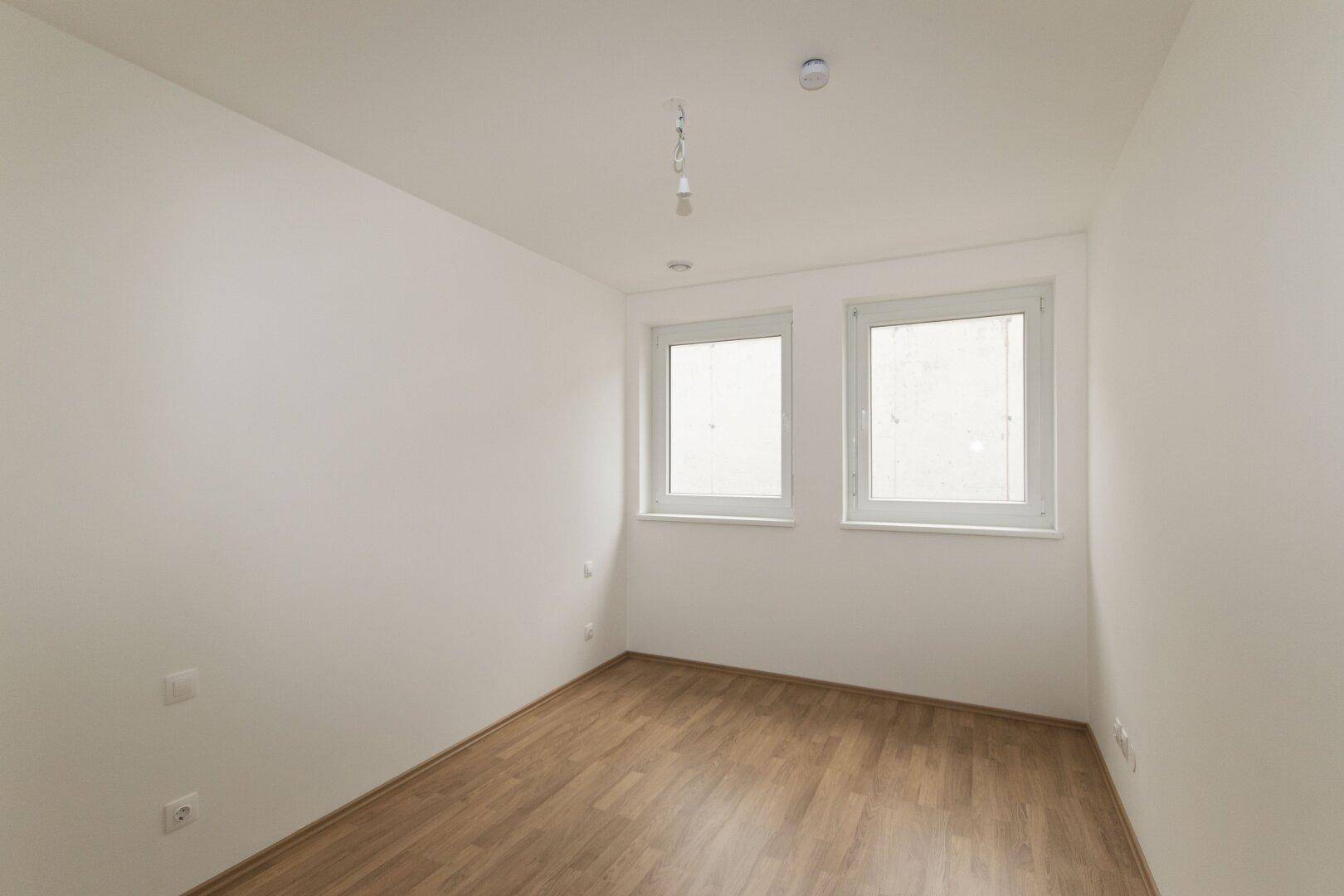 Unmöbliertes Schlafzimmer mit Holzboden und zwei Fenstern für viel Tageslicht.