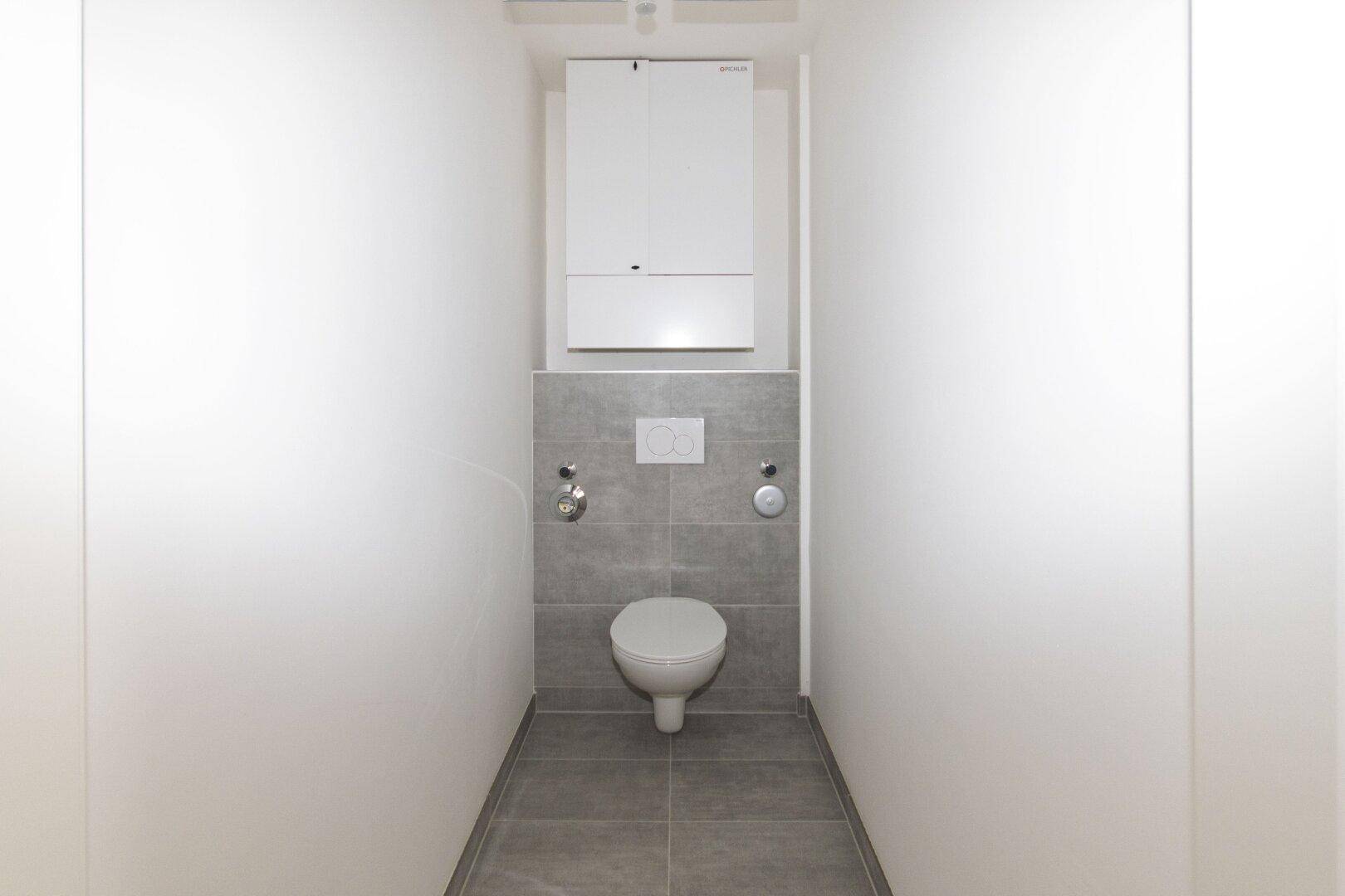 Separates WC mit wandhängender Toilette und grauen Bodenfliesen.