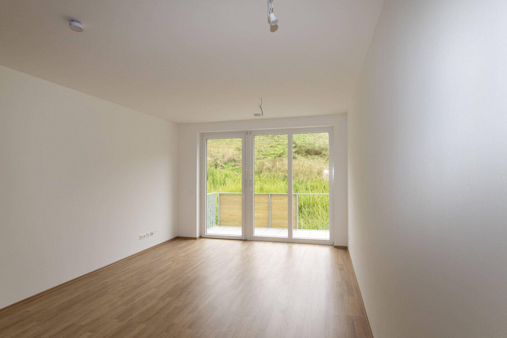 Heller Raum mit Holzboden und Zugang zu einem kleinen Balkon mit Blick ins Grüne.