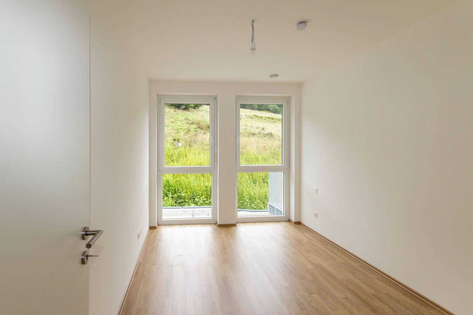 Helles Schlafzimmer mit Holzboden und großem Fenster mit Blick auf die grüne Landschaft.