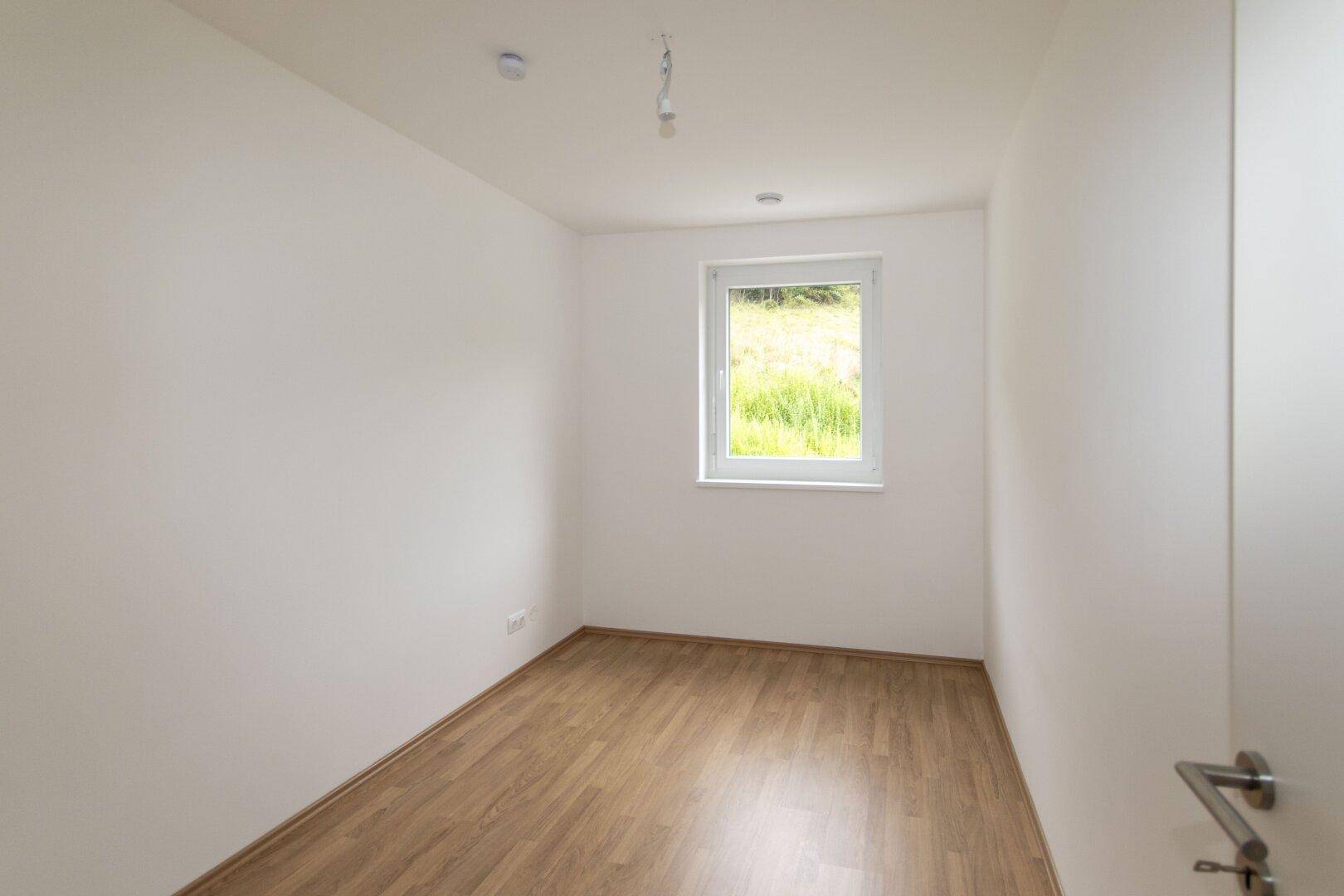 Kleinerer Raum mit hellem Holzboden und einem Fenster mit Ausblick ins Grüne.