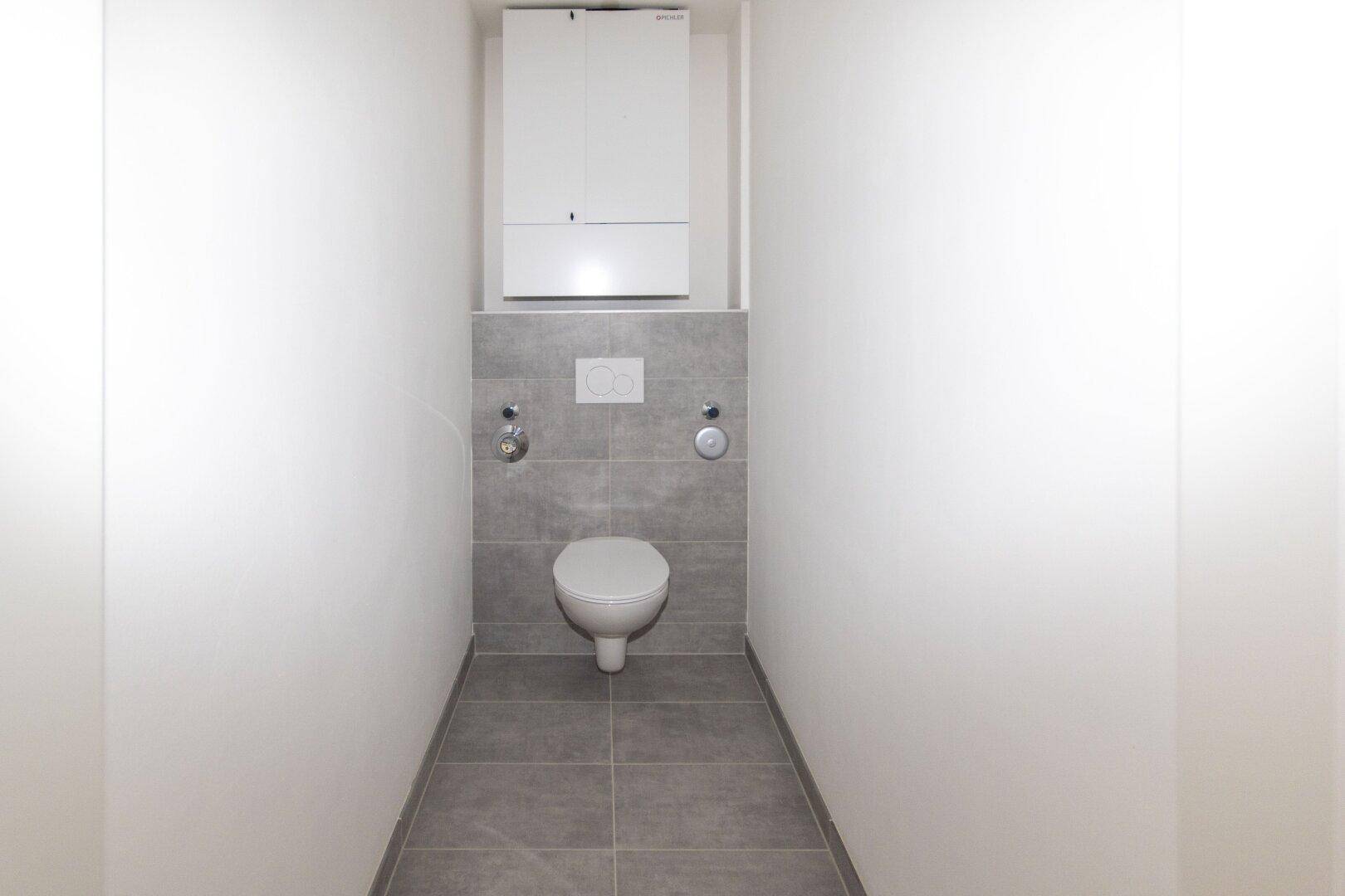 Separates WC mit wandhängender Toilette, grauen Fliesen und einem Spülkastenmodul.