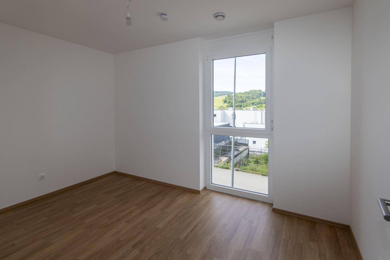 Helles Schlafzimmer mit Laminatboden und einem großen Fenster, das den Blick nach draußen freigibt.