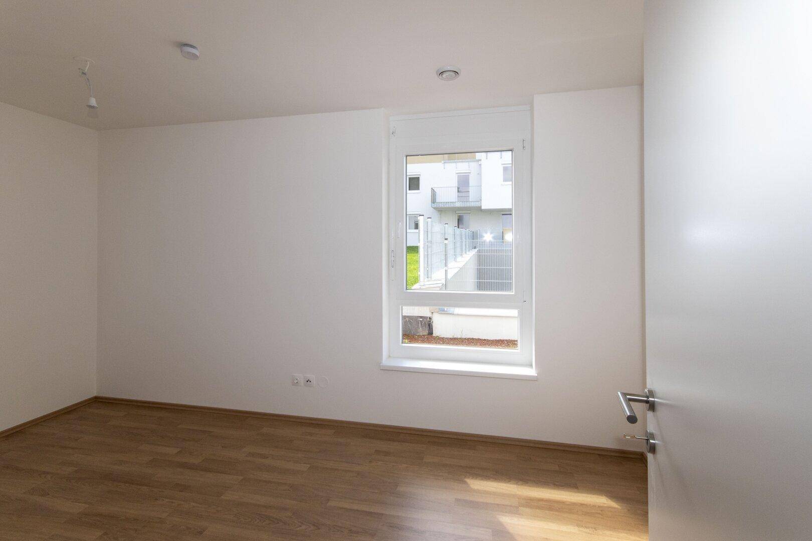 Leeres Schlafzimmer mit Laminatboden und einem Fenster, das auf den Innenhof blickt.