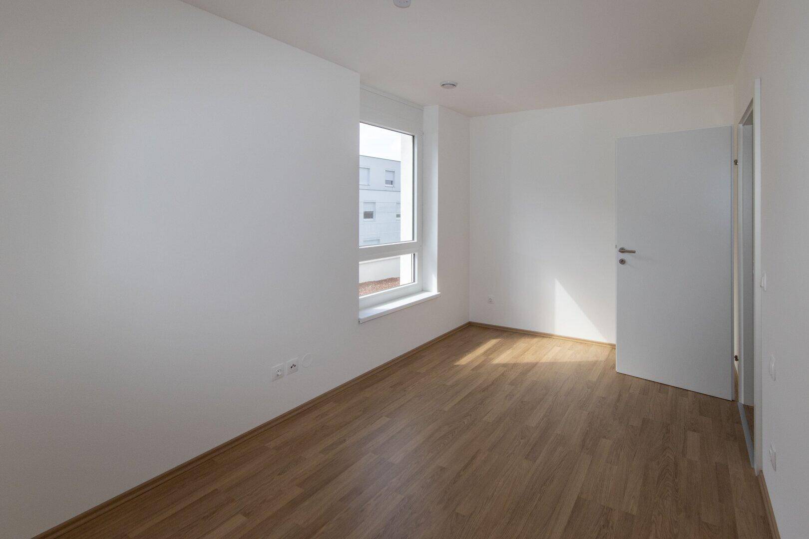 Heller Raum mit Laminatboden und einem Fenster, das viel Sonnenlicht in den Raum lässt.