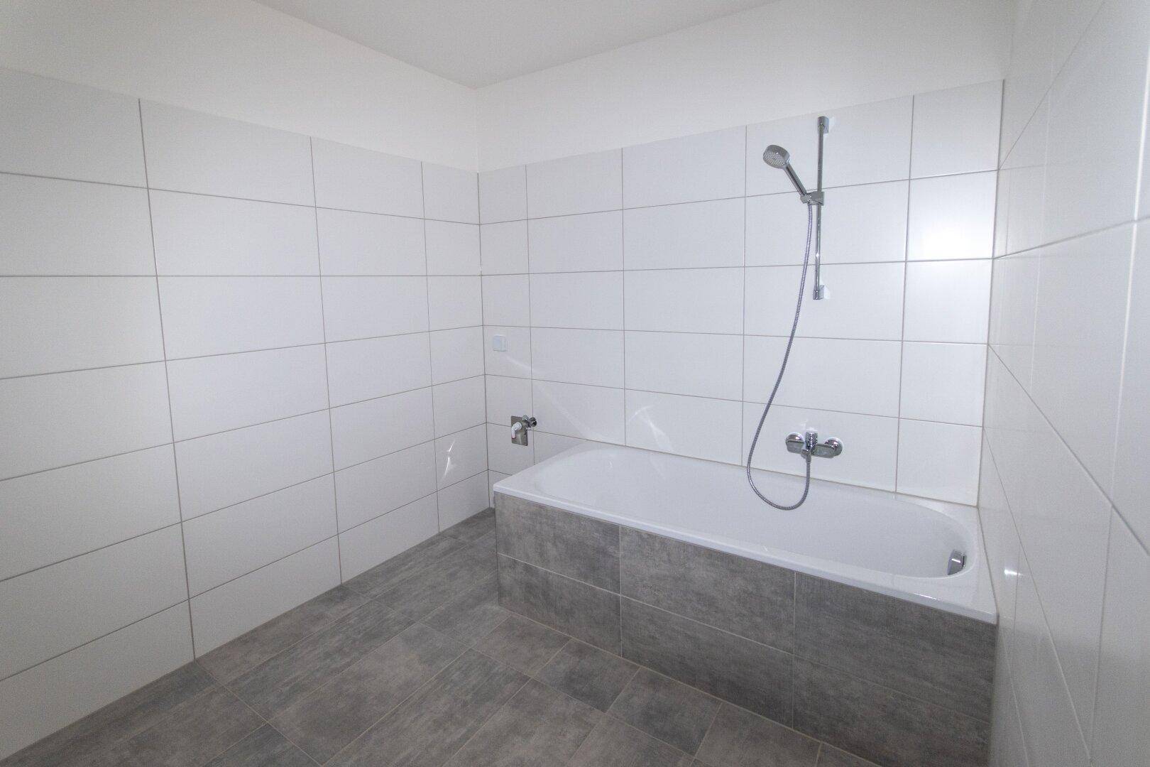 Modernes Badezimmer mit Badewanne und weißen Fliesen an den Wänden und grauen Bodenfliesen.